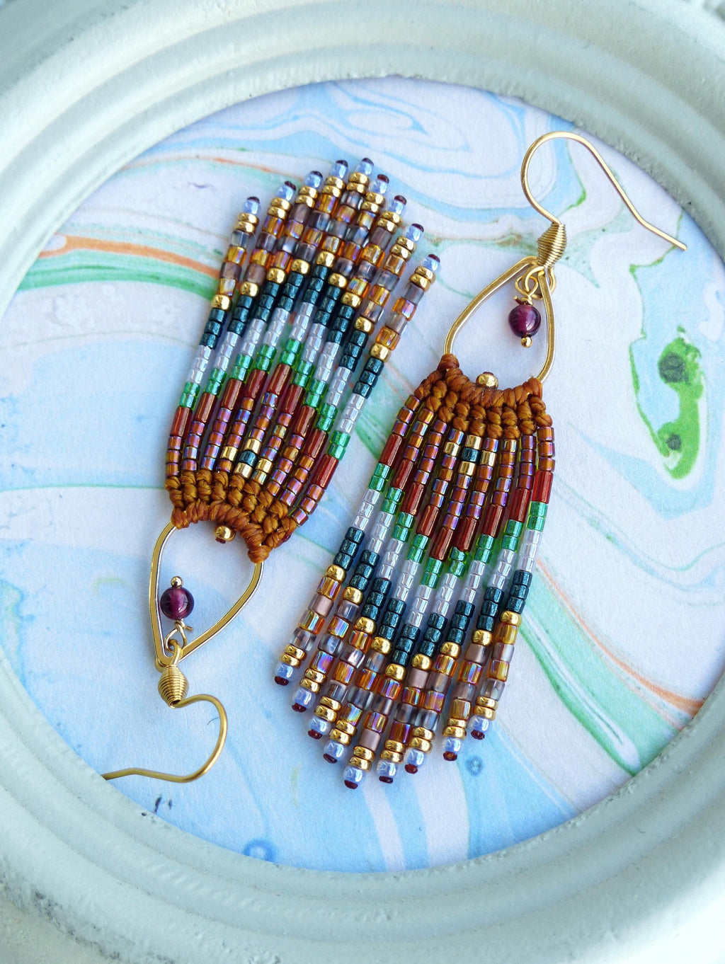 Peut inclure: Une paire de longues boucles d'oreilles pendantes en perles avec un schéma de couleurs brun, or, vert et violet. Les boucles d'oreilles ont un design en forme de larme avec une petite pierre précieuse violette foncée en haut.