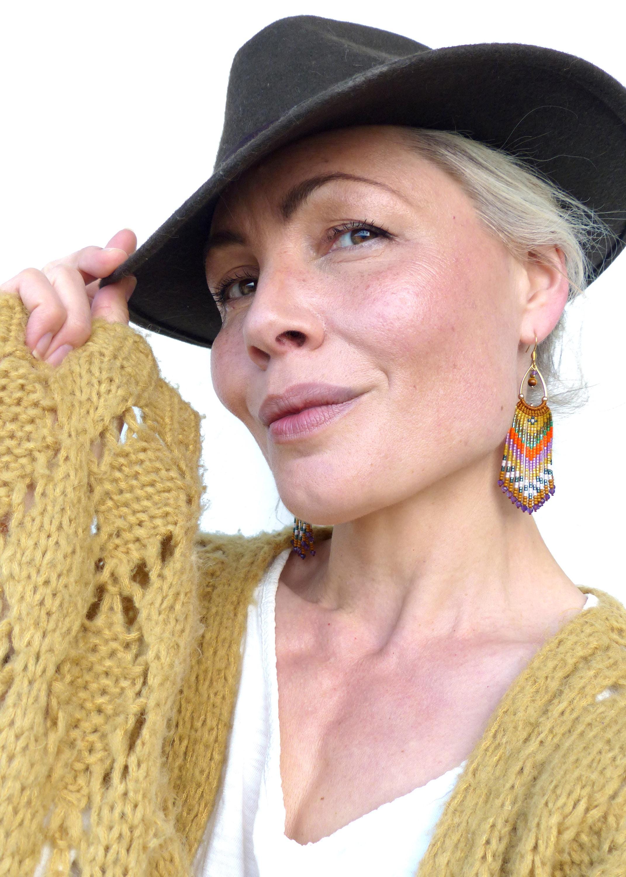 Peut inclure: Une femme portant un chapeau marron et un cardigan en tricot jaune avec une chemise blanche en dessous. Elle porte de longues boucles d'oreilles en perles colorées.