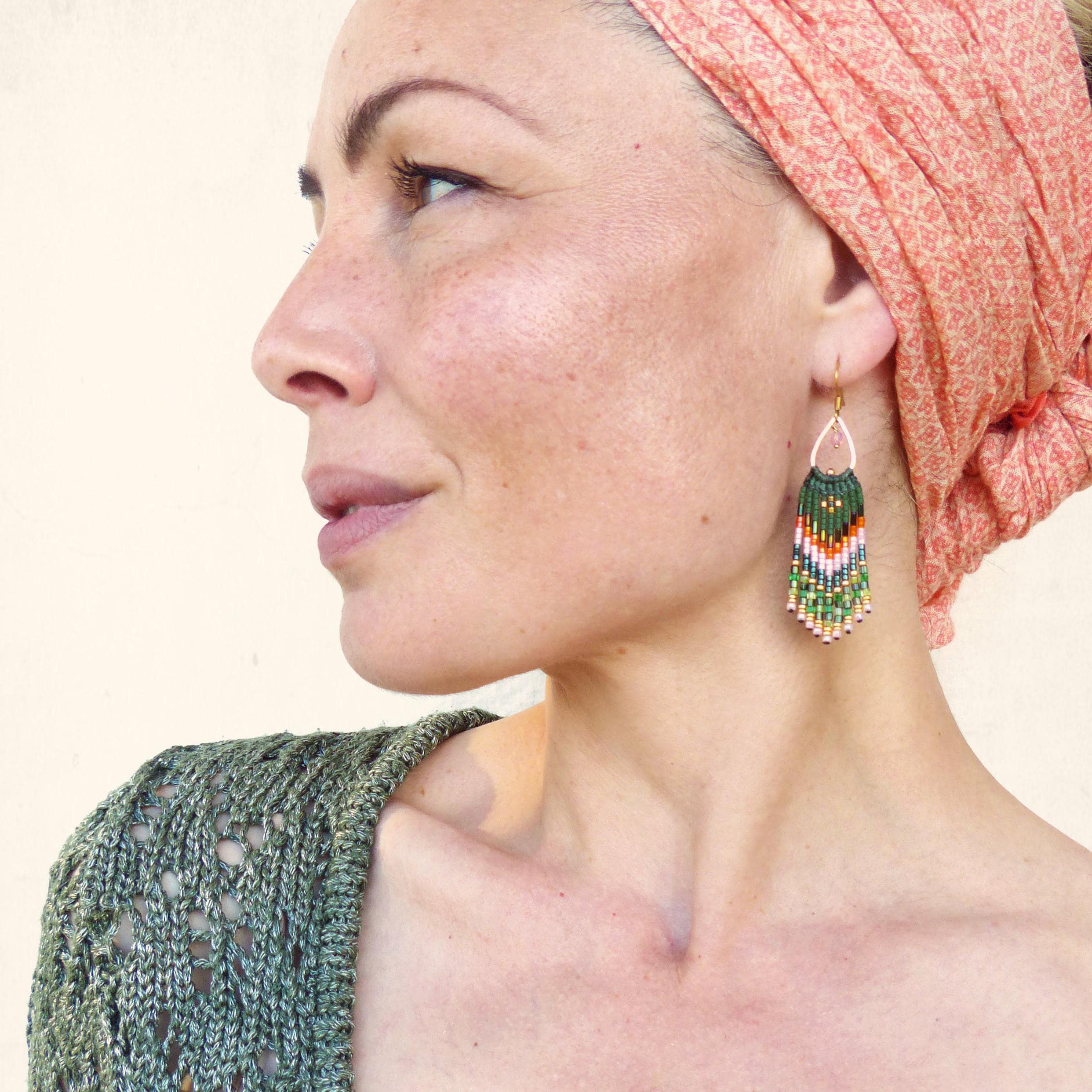 Peut inclure: Une paire de boucles d'oreilles pendantes en perles vertes, blanches et orange avec des accents dorés. Les boucles d'oreilles sont portées par une personne avec un foulard de tête couleur pêche.