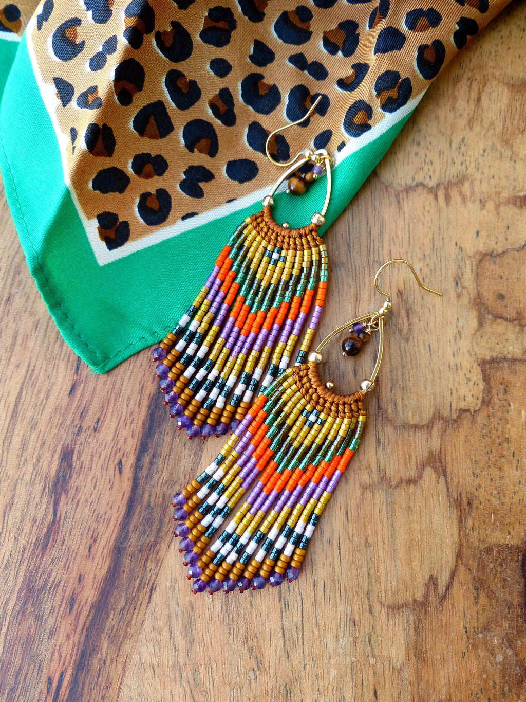 Peut inclure: Une paire de longues boucles d'oreilles en perles colorées avec des cercles dorés et des pierres œil de tigre. Les boucles d'oreilles présentent une variété de couleurs, notamment le violet, l'orange, le vert et l'or.