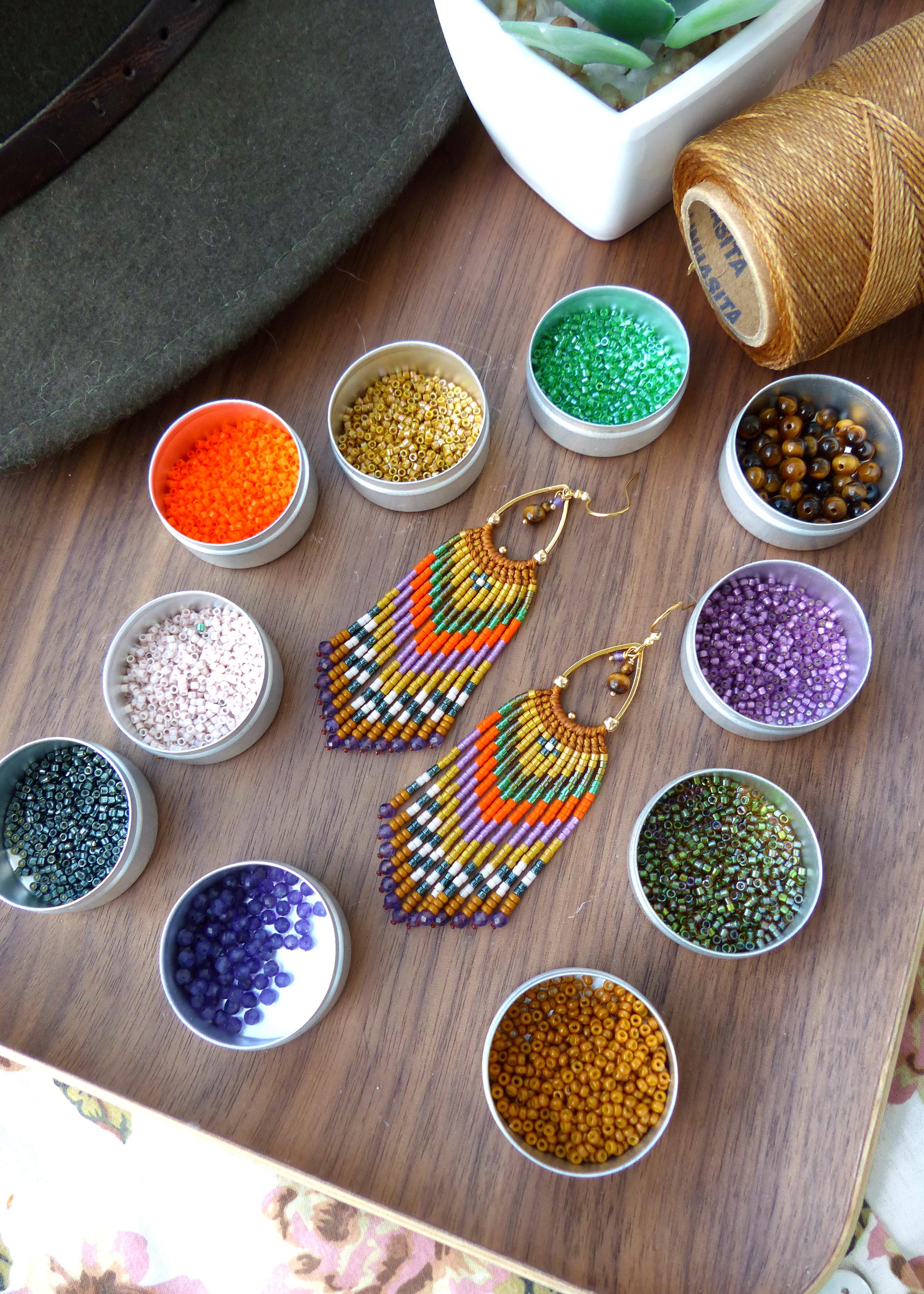 Peut inclure: Une paire de longues boucles d'oreilles en perles colorées avec un motif géométrique. Les boucles d'oreilles sont faites avec une variété de perles dans des tons d'orange, de vert, de violet, d'or et de blanc. Les boucles d'oreilles sont suspendues à des crochets dorés. L'image montre également une variété de perles dans de petits contenants.