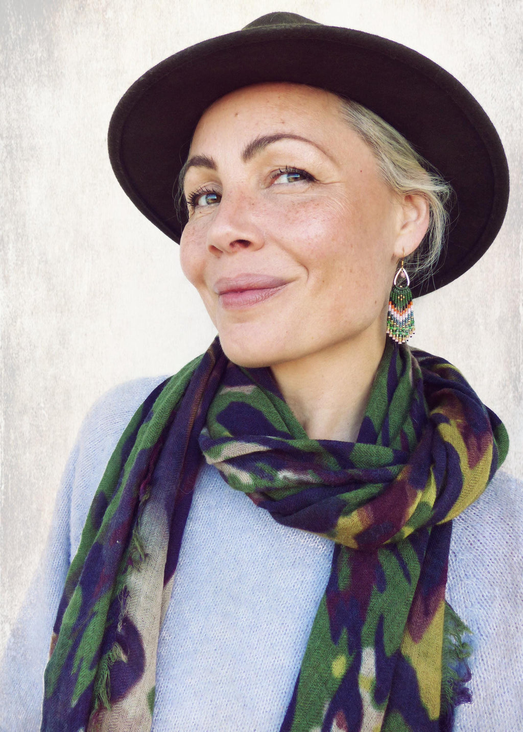 Peut inclure: Une femme portant un pull bleu avec une écharpe de camouflage vert et violet. Elle porte un chapeau noir et des boucles d'oreilles en perles vertes et blanches.