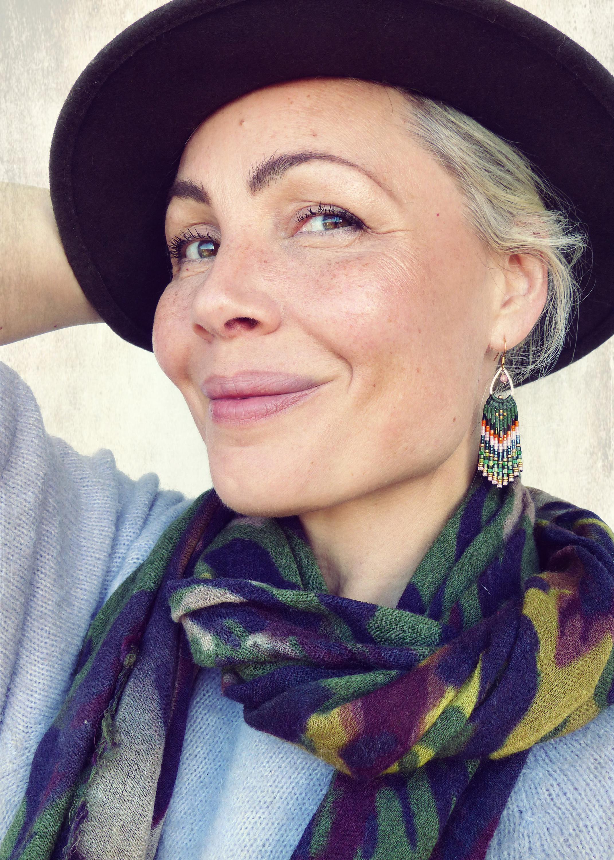 Peut inclure: Une femme portant un chapeau noir et une écharpe à motifs verts, bruns et blancs. Elle porte de longues boucles d'oreilles en perles vertes, orange et blanches.