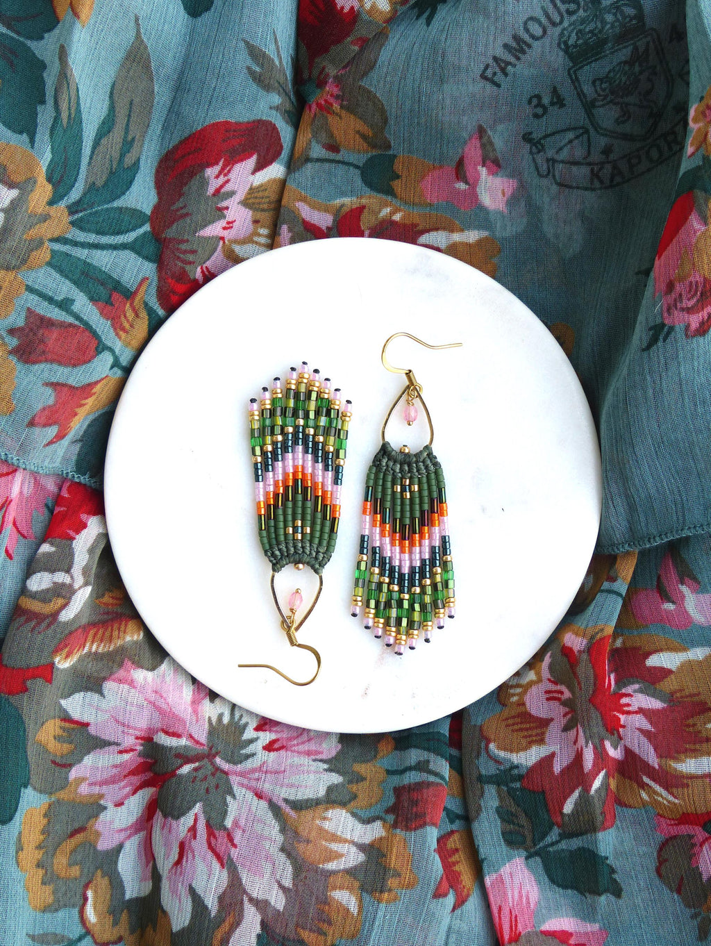 Peut inclure: Une paire de boucles d'oreilles pendantes en perles vertes, roses et dorées avec un accent de pierre rose. Les boucles d'oreilles sont sur une assiette blanche.