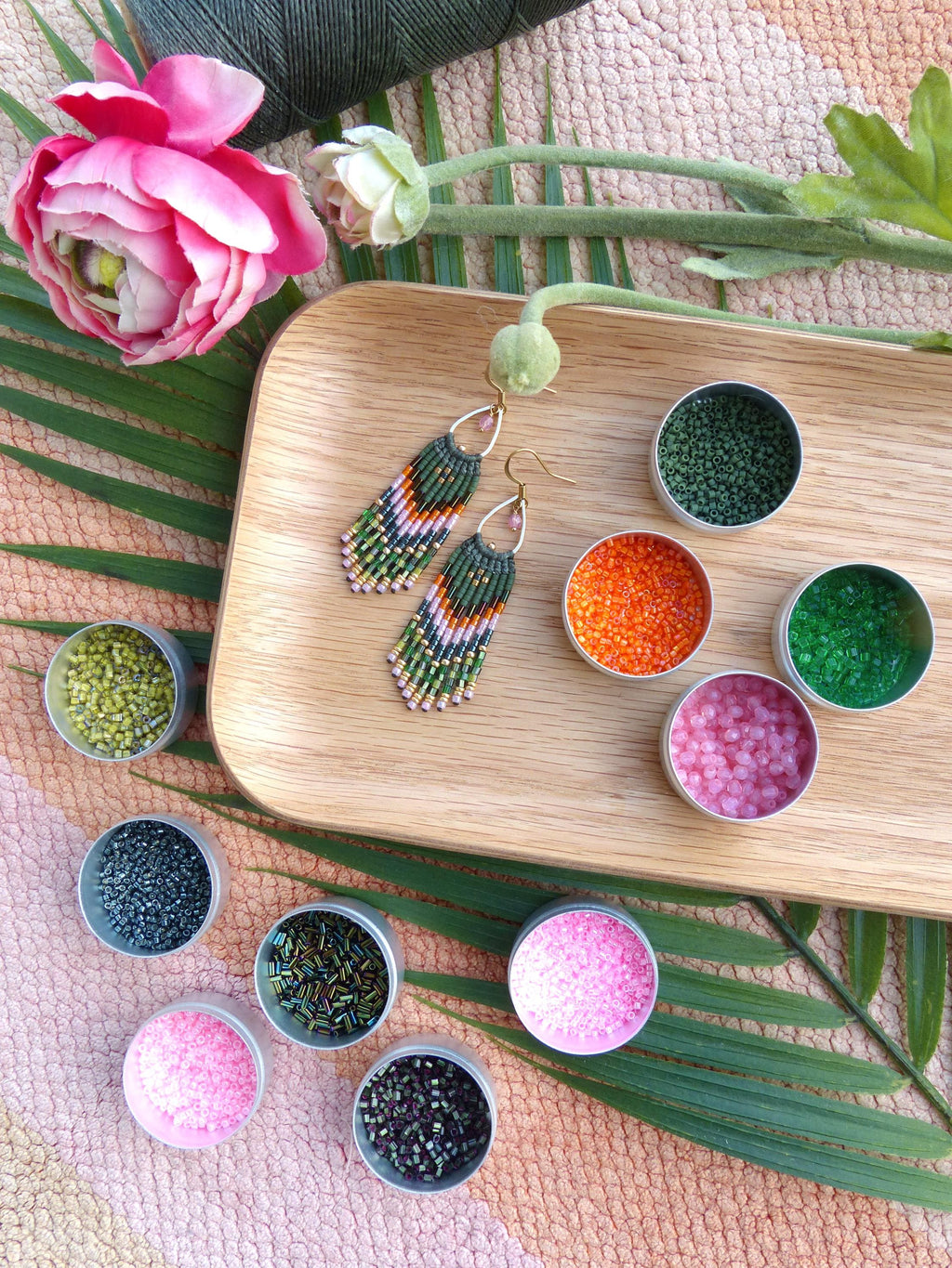 Peut inclure: Une paire de boucles d'oreilles en perles vertes, roses et dorées avec des franges, présentée sur un plateau en bois avec de petits récipients de perles de différentes couleurs.
