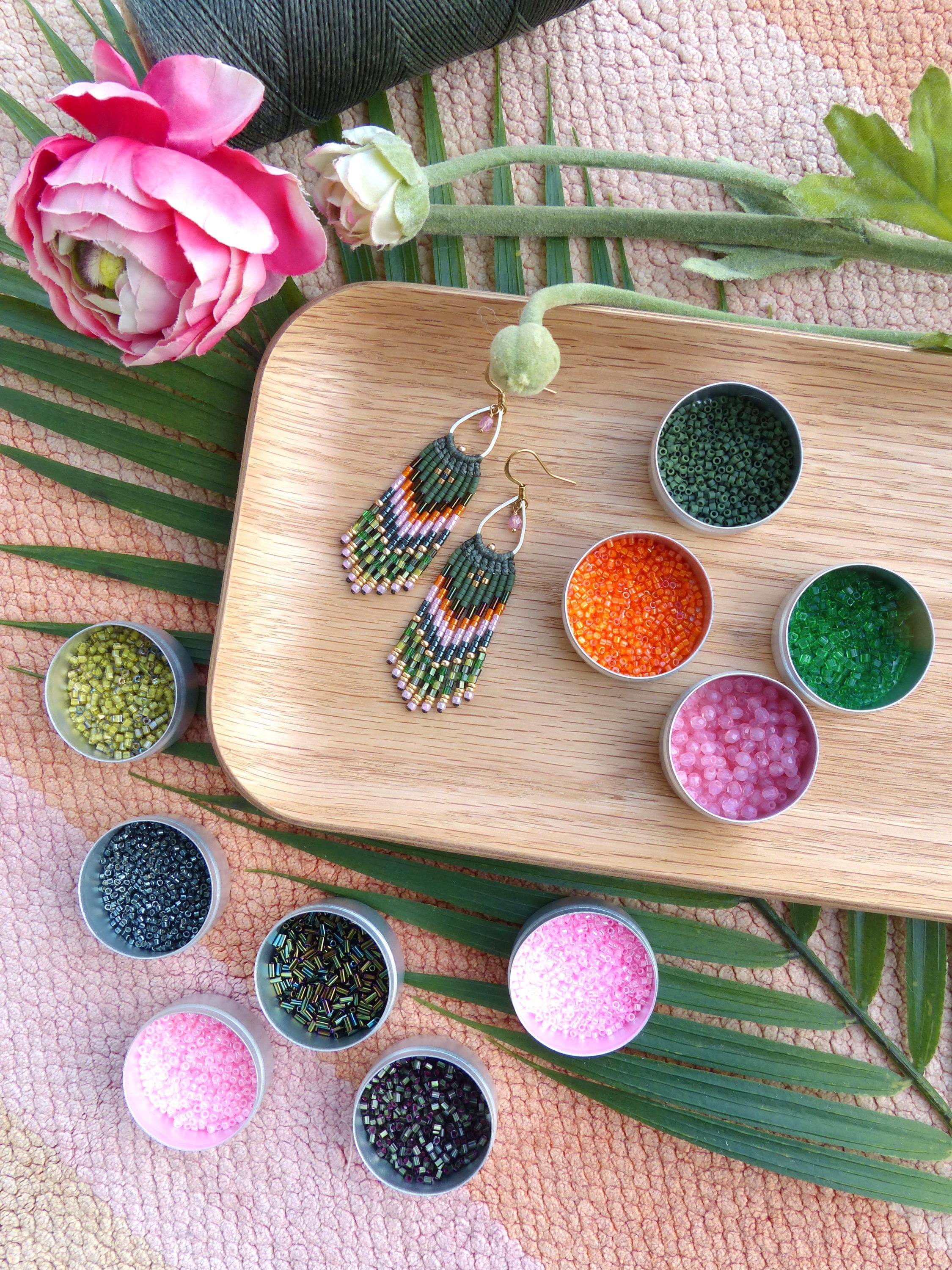 Peut inclure: Une paire de boucles d'oreilles en perles vertes, roses et dorées avec des franges, présentée sur un plateau en bois avec de petits récipients de perles de différentes couleurs.