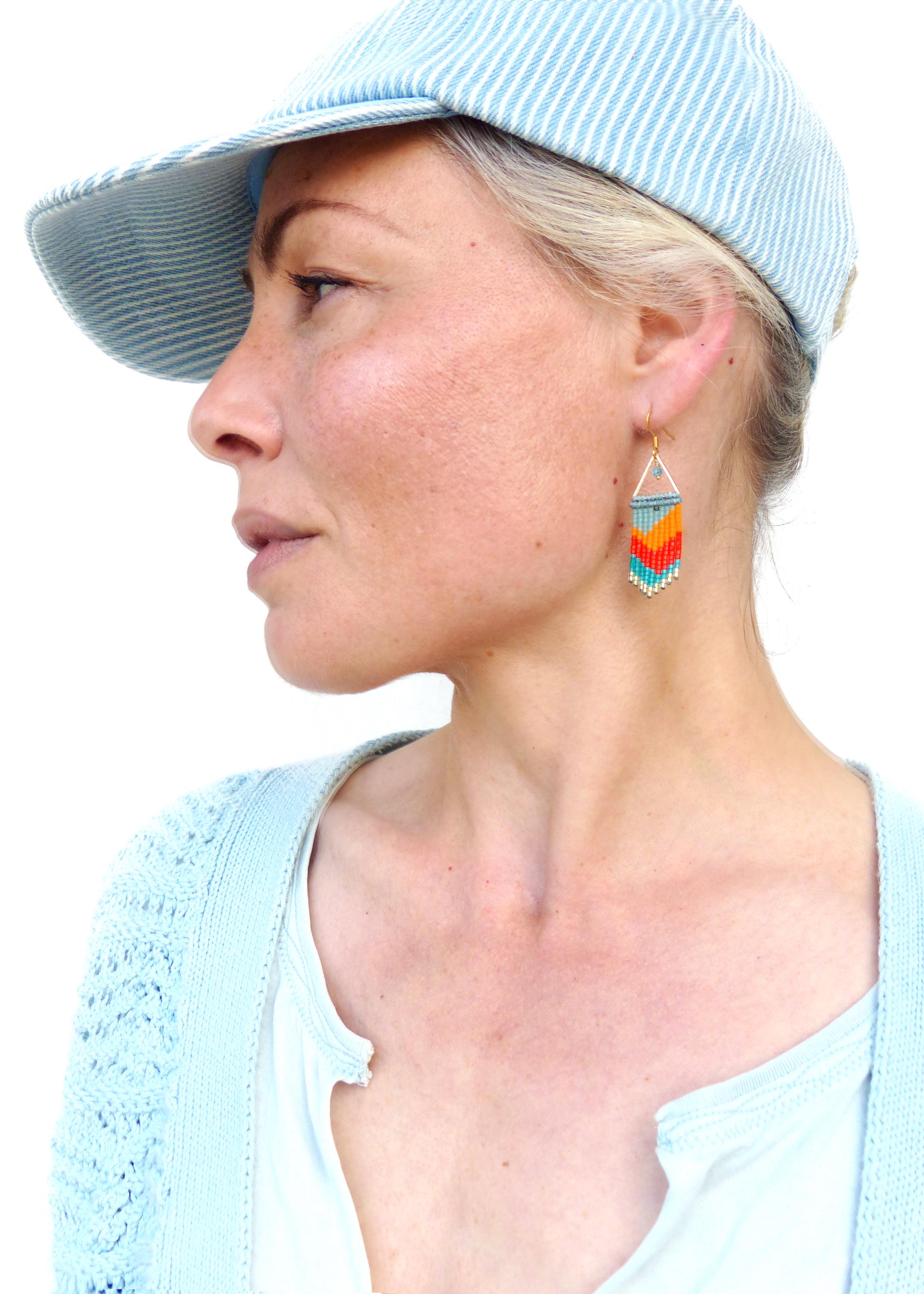 Peut inclure: Une femme portant une casquette de baseball rayée bleu clair et un cardigan en crochet bleu clair. Elle porte une paire de boucles d'oreilles en perles colorées avec un motif en chevron.