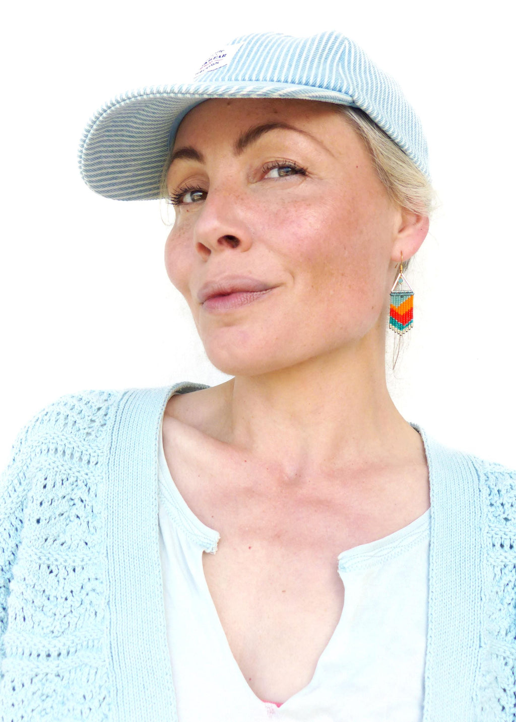 Peut inclure: Une femme portant une casquette de baseball rayée bleu clair avec un cardigan en crochet blanc et un t-shirt blanc. Elle porte des boucles d'oreilles en perles colorées.
