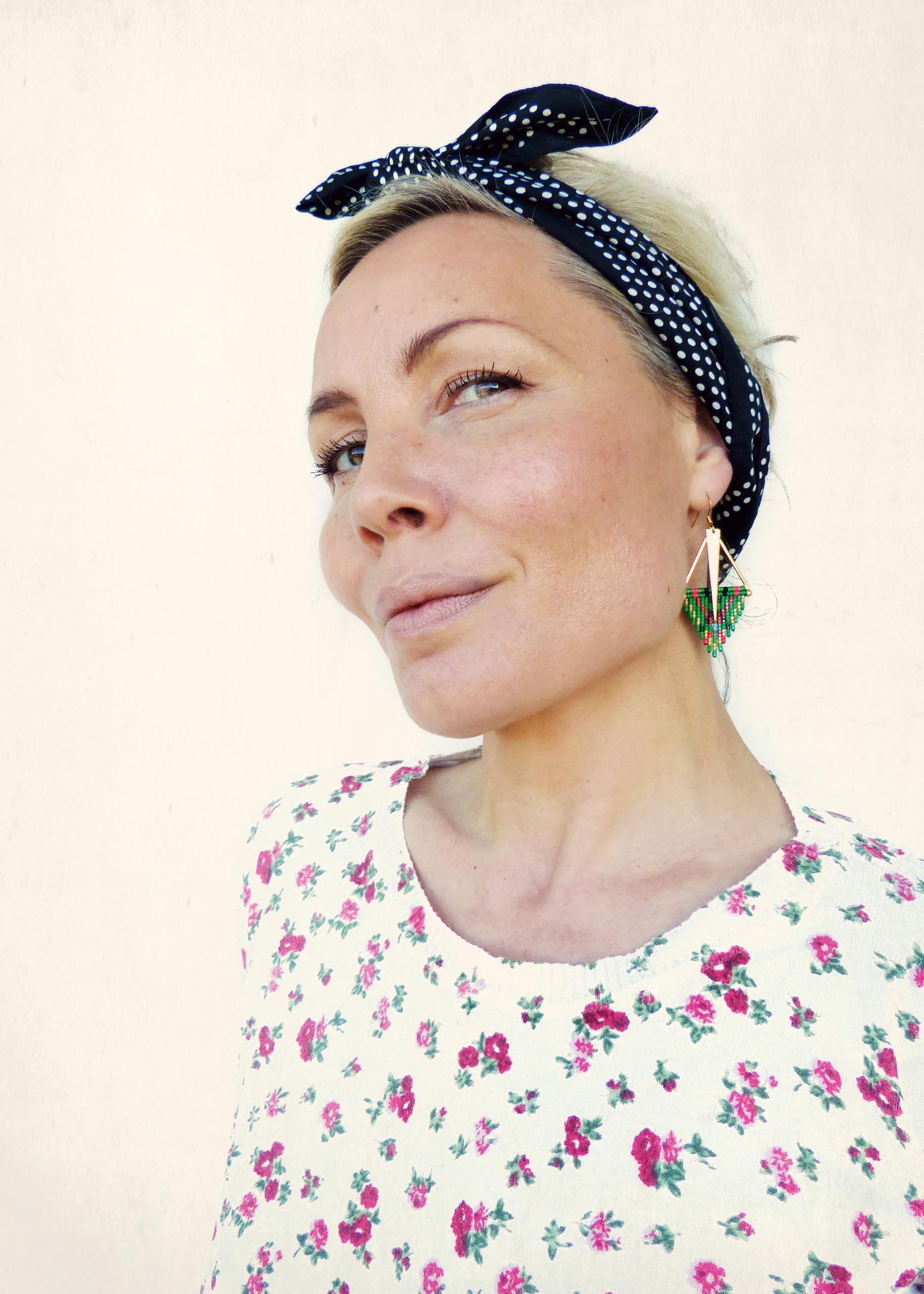 Peut inclure: Une femme aux cheveux blonds portant un bandeau à pois noirs et blancs et des boucles d'oreilles en perles vertes et dorées. Elle porte une blouse blanche à motif floral.
