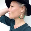 Peut inclure: Boucles d'oreilles créoles dorées avec des détails de perles vertes et noires. Une perle rouge et un charm étoile dorée pendent de la créole. La personne porte un chapeau noir et un haut vert foncé.