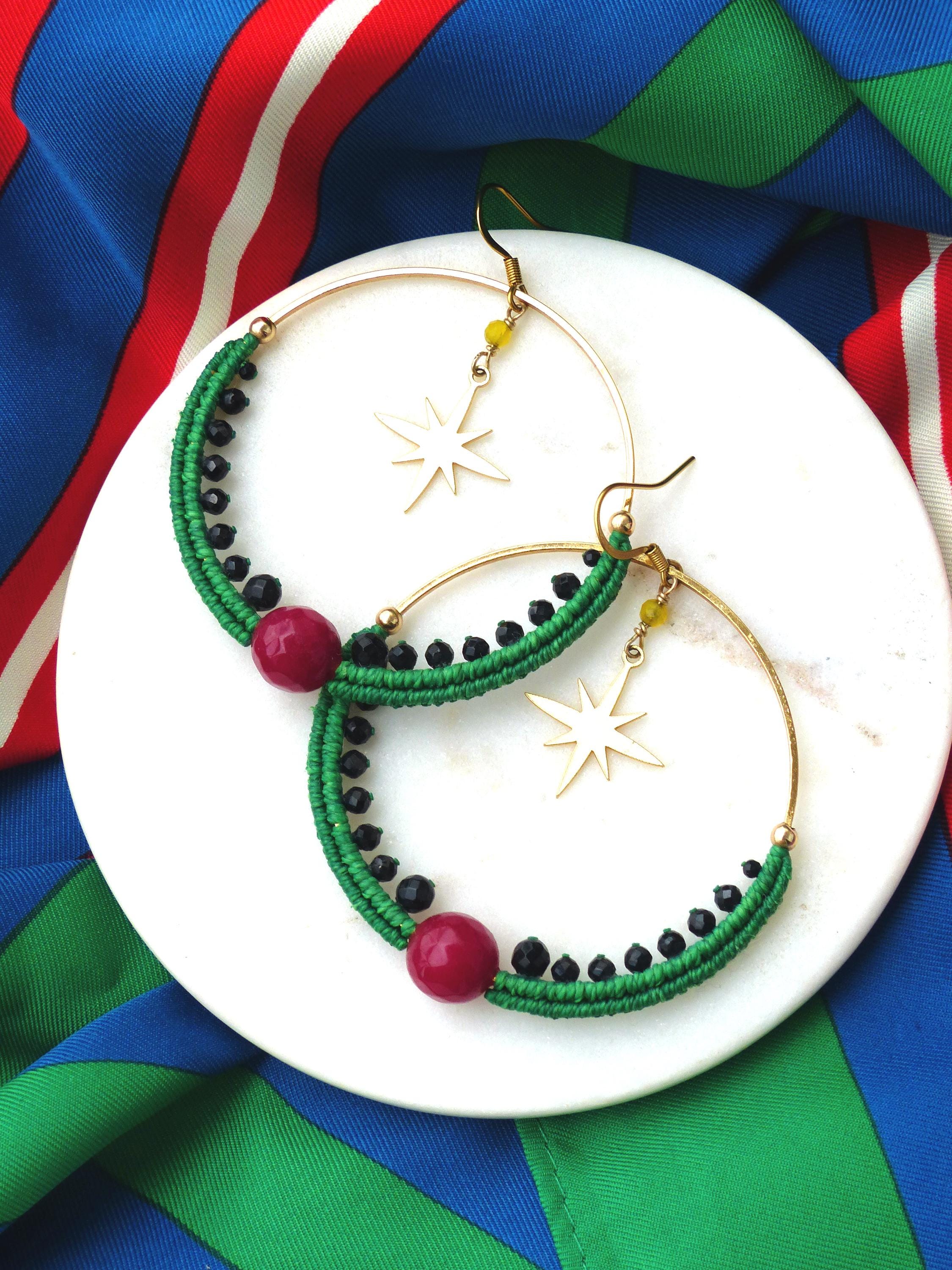 Peut inclure: Paire de boucles d'oreilles créoles dorées avec des détails en macramé vert, des perles noires et une grosse perle rouge. Chaque boucle d'oreille comporte un charm en forme d'étoile dorée et des perles jaunes. Présentées sur une surface blanche.