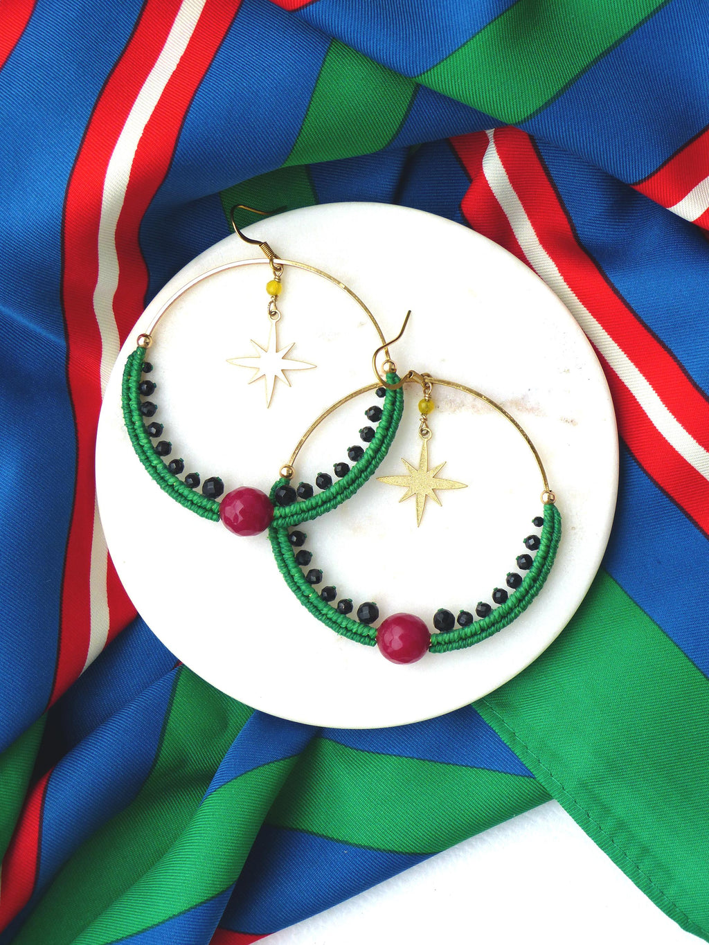 Peut inclure: Boucles d'oreilles créoles dorées avec détails en macramé vert, perles noires et une grosse perle rouge. Chaque boucle d'oreille a un charm en forme d'étoile dorée et de petites perles jaunes. Les boucles d'oreilles reposent sur une assiette blanche.
