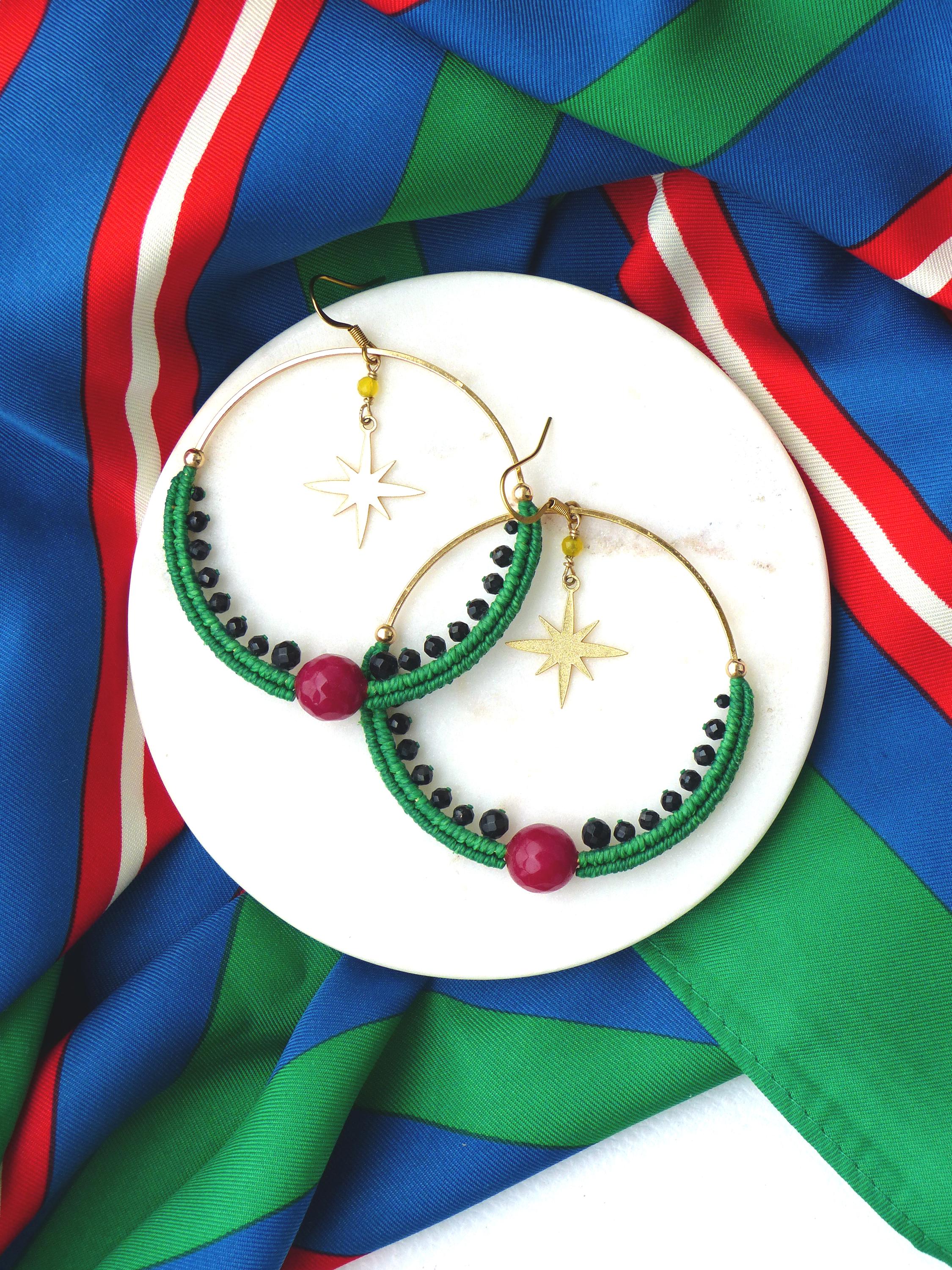Peut inclure: Boucles d'oreilles créoles dorées avec détails en macramé vert, perles noires et une grosse perle rouge. Chaque boucle d'oreille a un charm en forme d'étoile dorée et de petites perles jaunes. Les boucles d'oreilles reposent sur une assiette blanche.