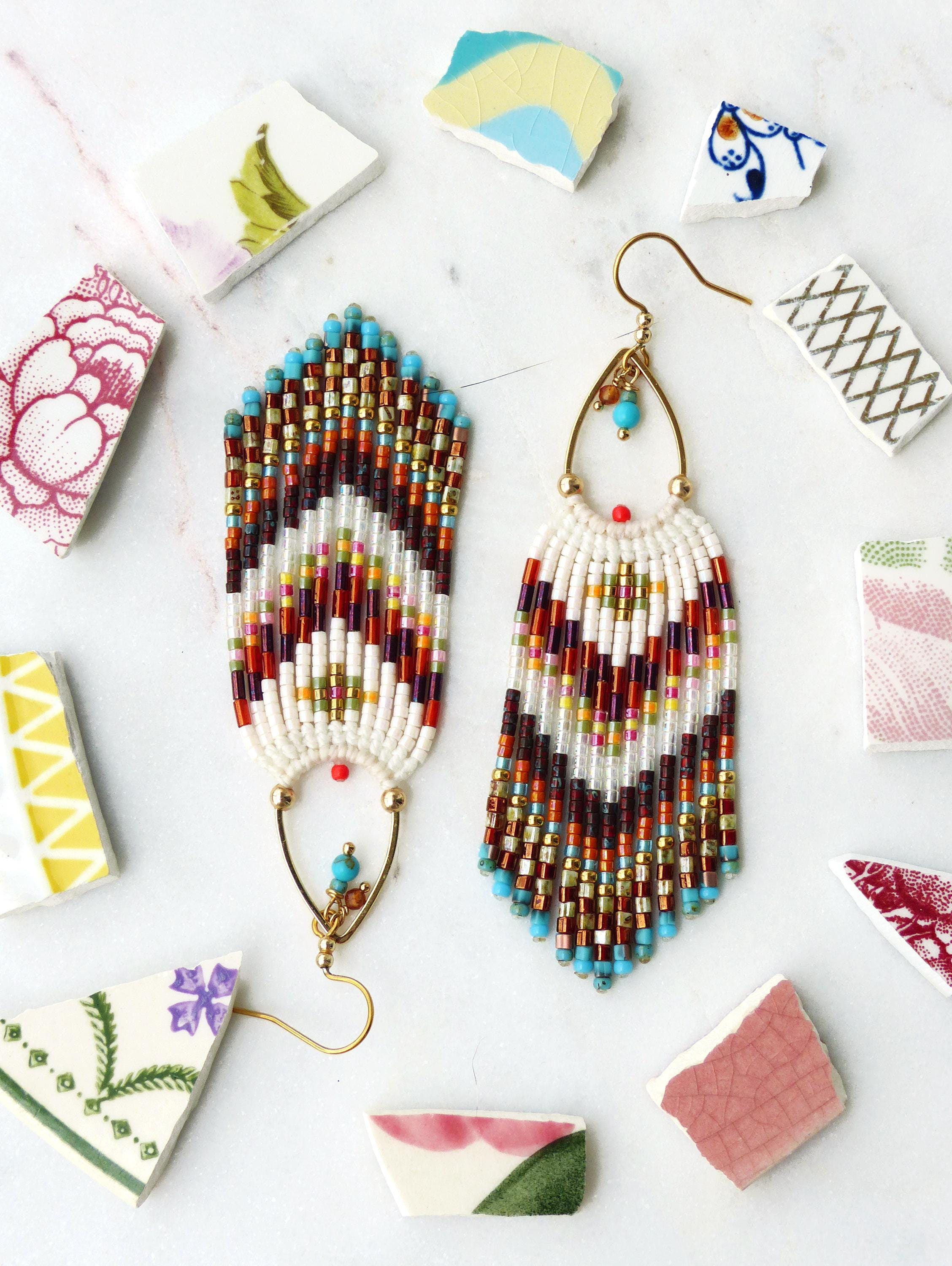 Peut inclure: Paire de boucles d'oreilles perlées faites à la main, à base blanche et motif chevron coloré rouge, marron et or. Les boucles d'oreilles ont des crochets d'oreilles dorés et des accents turquoise. Des éclats de céramique décorent l'image.