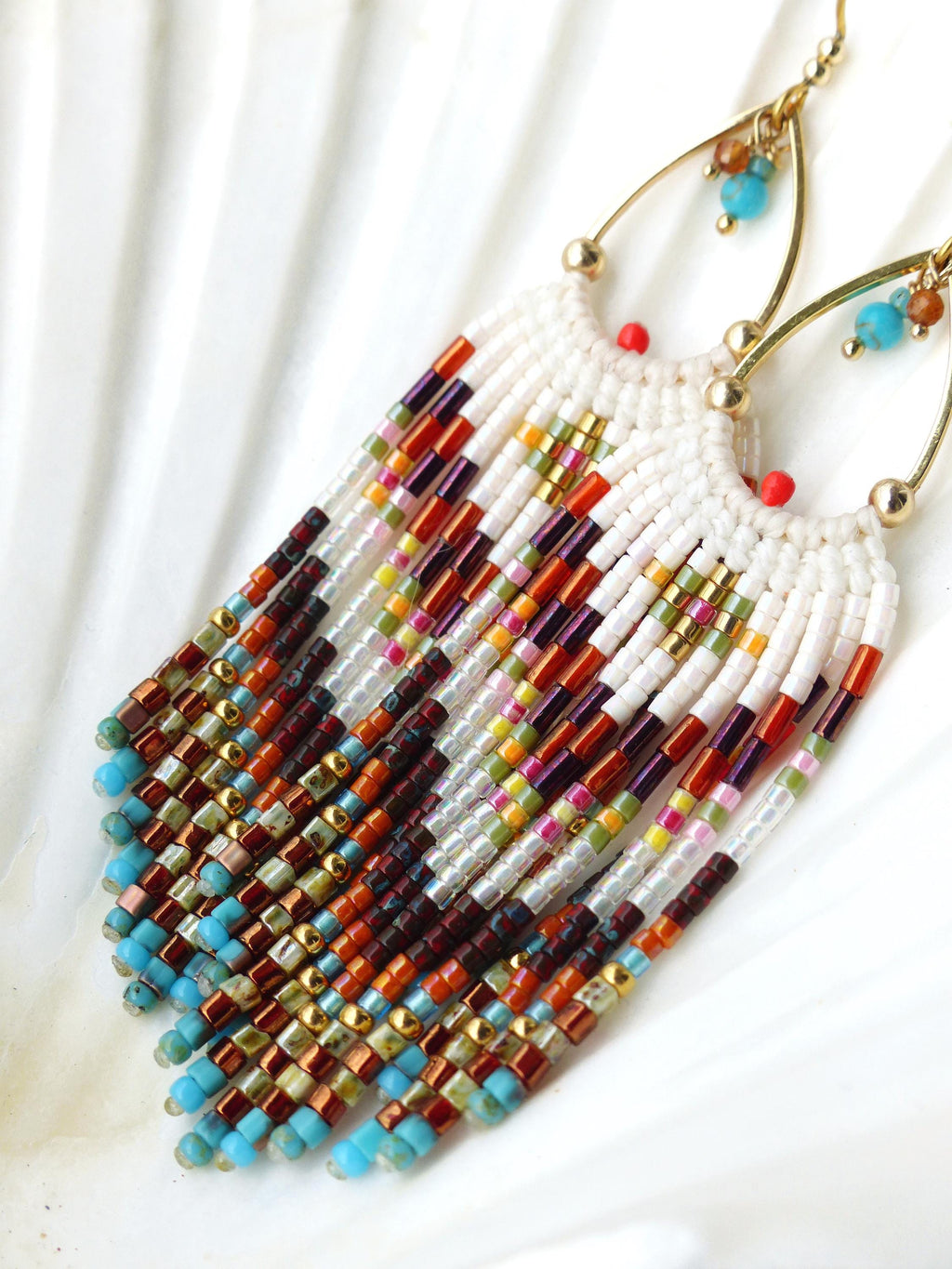 Peut inclure: Boucles d'oreilles perlées faites à la main avec une base blanche et des franges colorées. Les boucles d'oreilles présentent un mélange de perles rouges, oranges, violettes et turquoise, avec des accents dorés. Style bohème unique.