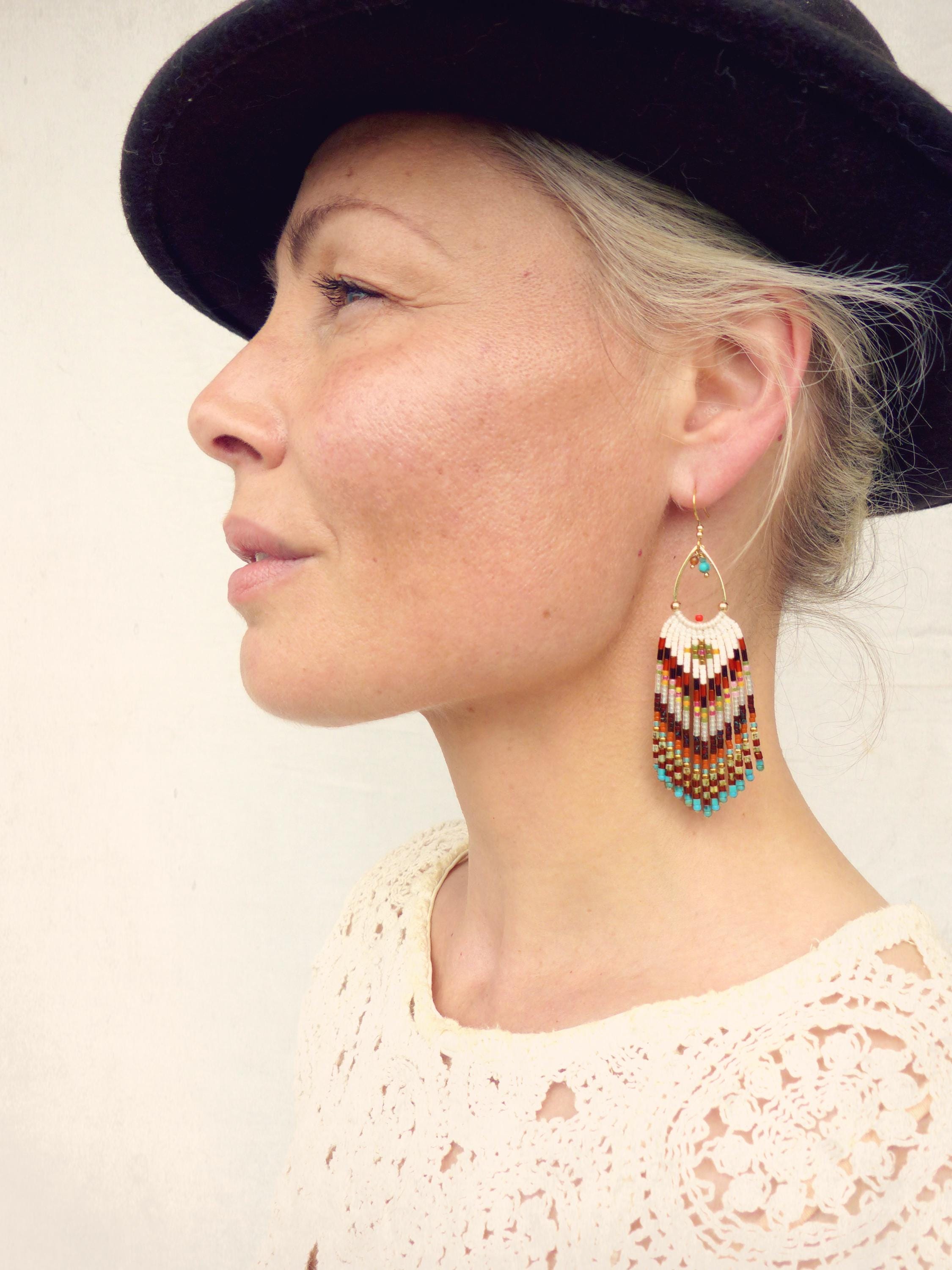 Peut inclure: Boucles d'oreilles en or ornées de perles avec un motif en forme de goutte d'eau. Les boucles d'oreilles présentent une frange de perles colorées dans les tons blanc, rouge, marron et turquoise. La personne porte un chapeau noir et un haut crème.