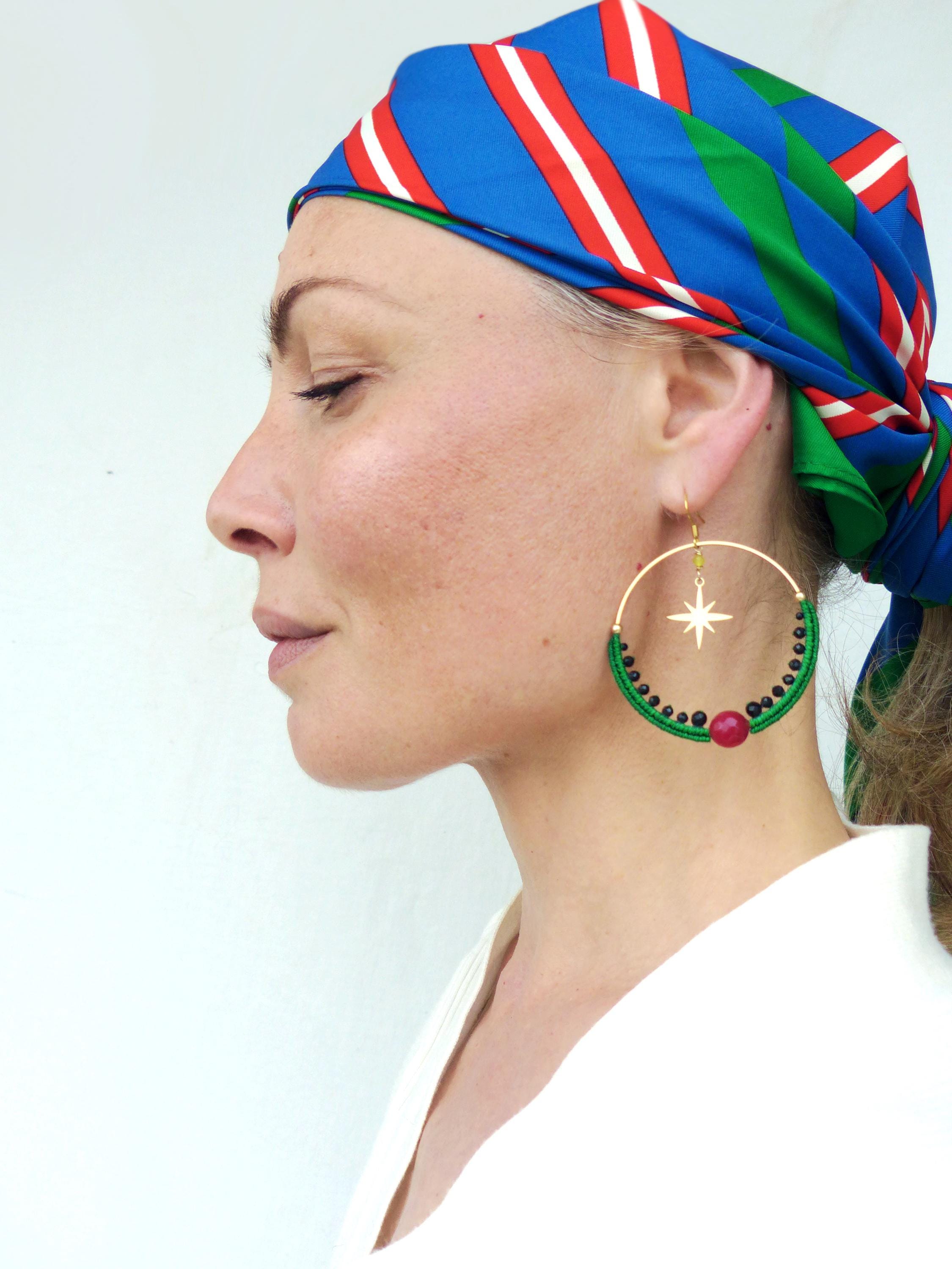 Peut inclure: Boucles d'oreilles créoles dorées avec un motif de perles vertes et noires, un charme en forme d'étoile et un accent de perle rouge. Les boucles d'oreilles sont portées avec un foulard à rayures bleues, rouges et blanches.