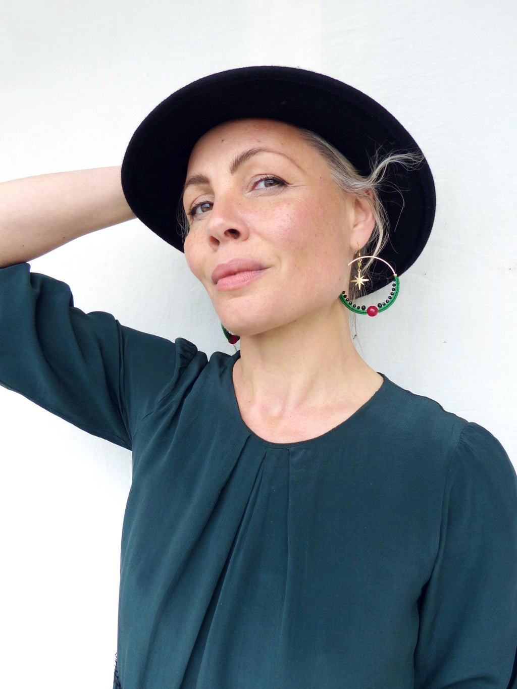 Peut inclure: Une personne portant un chapeau noir et un chemisier vert sarcelle foncé. Elle porte de grandes boucles d'oreilles créoles dorées avec un motif vert et rouge et une breloque étoile dorée. Les boucles d'oreilles sont un accessoire unique et accrocheur.
