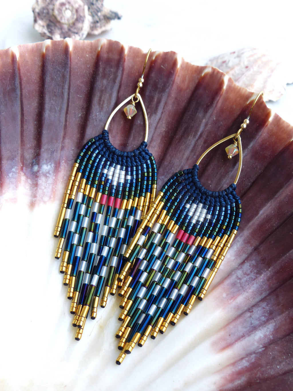 Peut inclure: Boucles d'oreilles faites à la main avec une partie supérieure bleu marine et une frange de perles dorées, bleues, argentées et rouges. Les boucles d'oreilles ont des crochets d'oreilles dorés et sont présentées sur un coquillage.