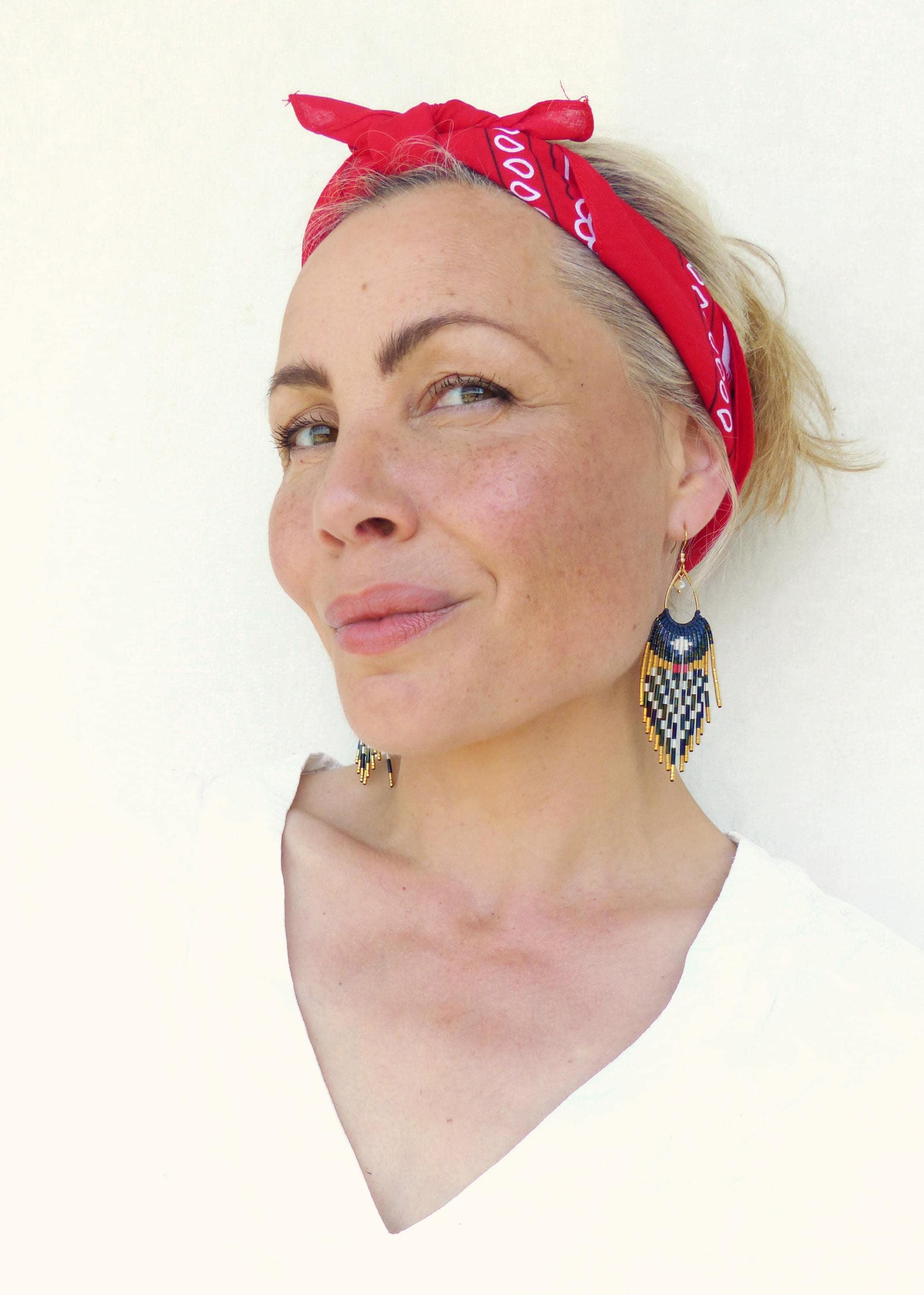Peut inclure: Une femme porte des boucles d'oreilles en perles avec un motif géométrique bleu, noir, blanc et or. Elle porte également un bandana rouge noué autour de la tête. Les boucles d'oreilles sont au centre de l'image.
