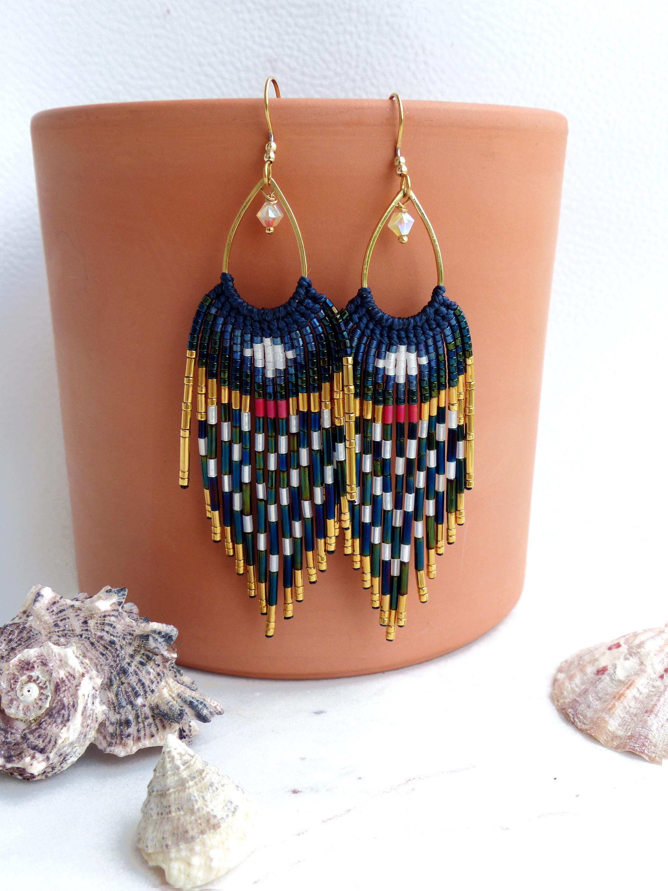 Peut inclure: Boucles d'oreilles macramé faites à la main, avec une section supérieure bleu marine et un motif géométrique blanc, rouge et vert. Des perles et des franges dorées complètent le design. Les boucles d'oreilles sont suspendues à des crochets d'oreilles dorés.