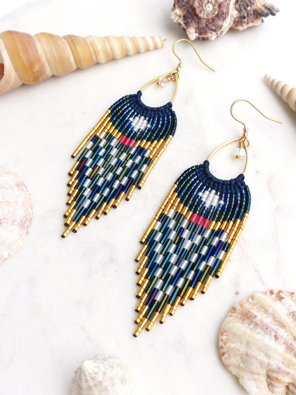 Peut inclure: Paire de boucles d'oreilles faites à la main avec une section supérieure bleu marine et une frange de perles dorées, argentées et bleues. Les boucles d'oreilles ont des crochets d'oreilles dorés et sont présentées sur une surface blanche avec des coquillages.