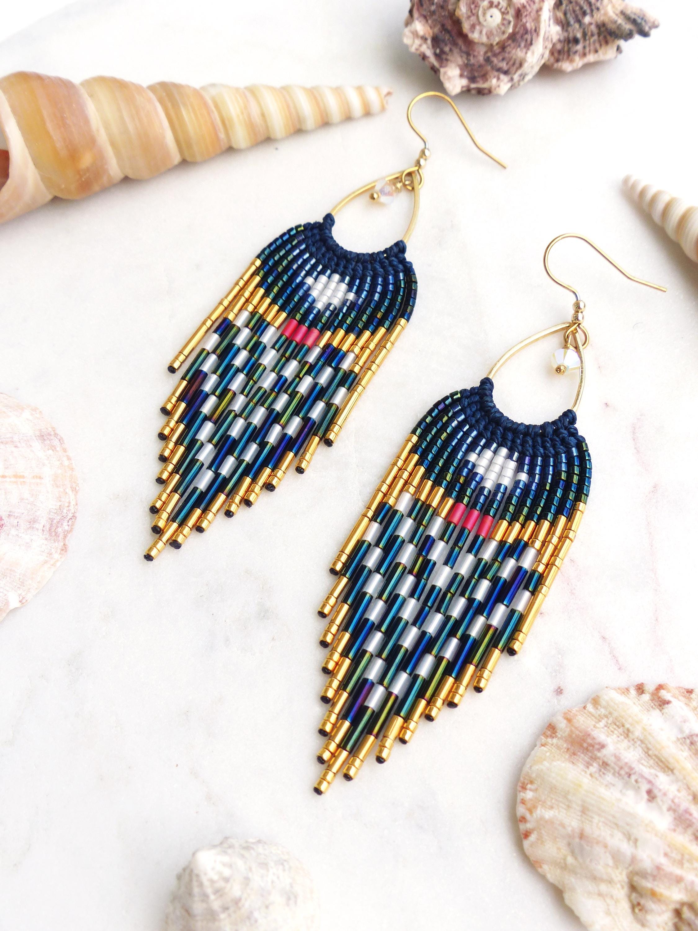 Peut inclure: Paire de boucles d'oreilles faites à la main avec une section supérieure bleu marine et une frange de perles dorées, argentées et bleues. Les boucles d'oreilles ont des crochets d'oreilles dorés et sont présentées sur une surface blanche avec des coquillages.