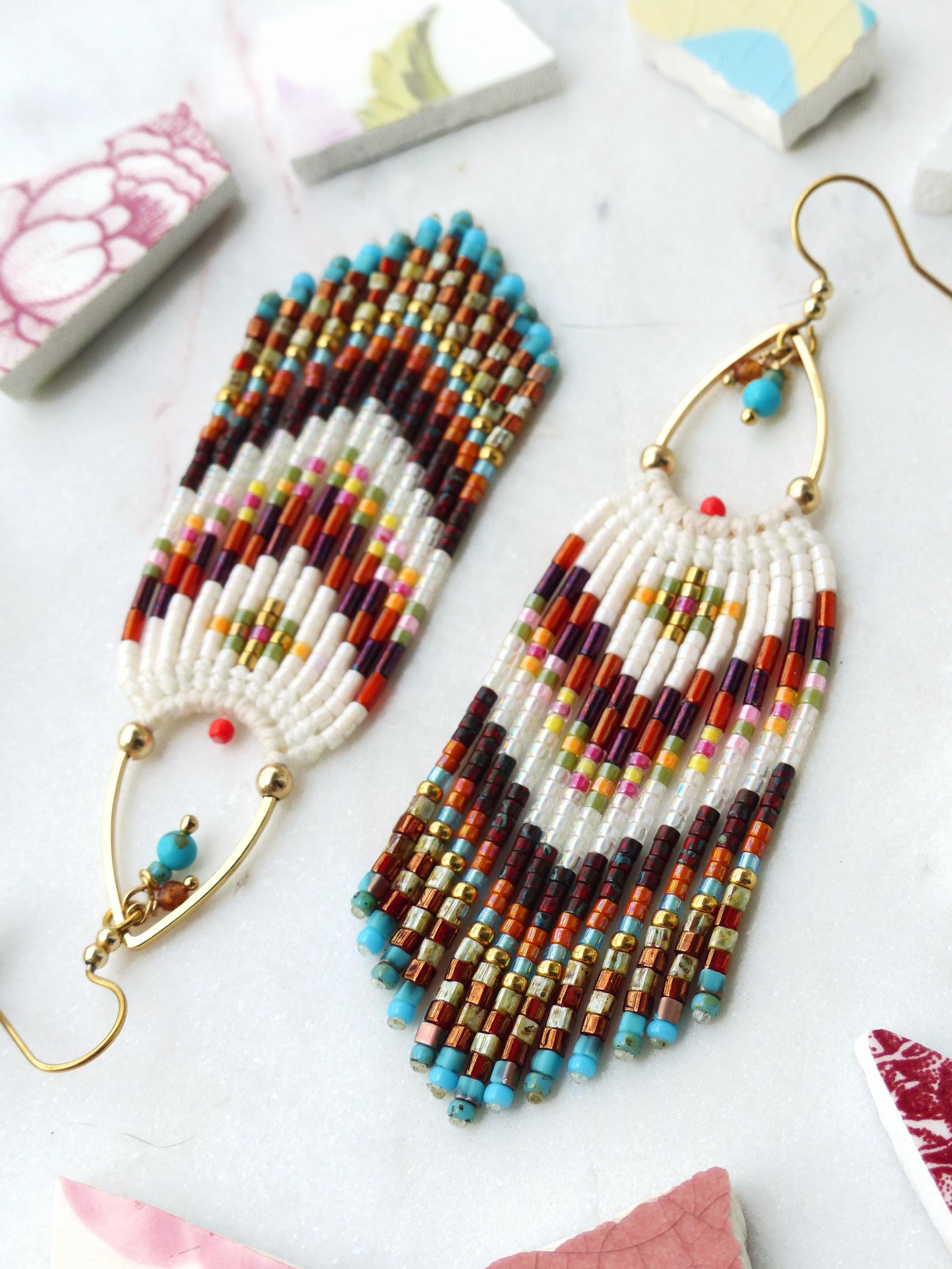 Peut inclure: Une paire de boucles d'oreilles faites à la main avec une base blanche et des accents colorés. Les boucles d'oreilles présentent un motif de franges avec des perles turquoise, dorées, marron et rouges. Les crochets d'oreilles dorés ont une forme de goutte d'eau.