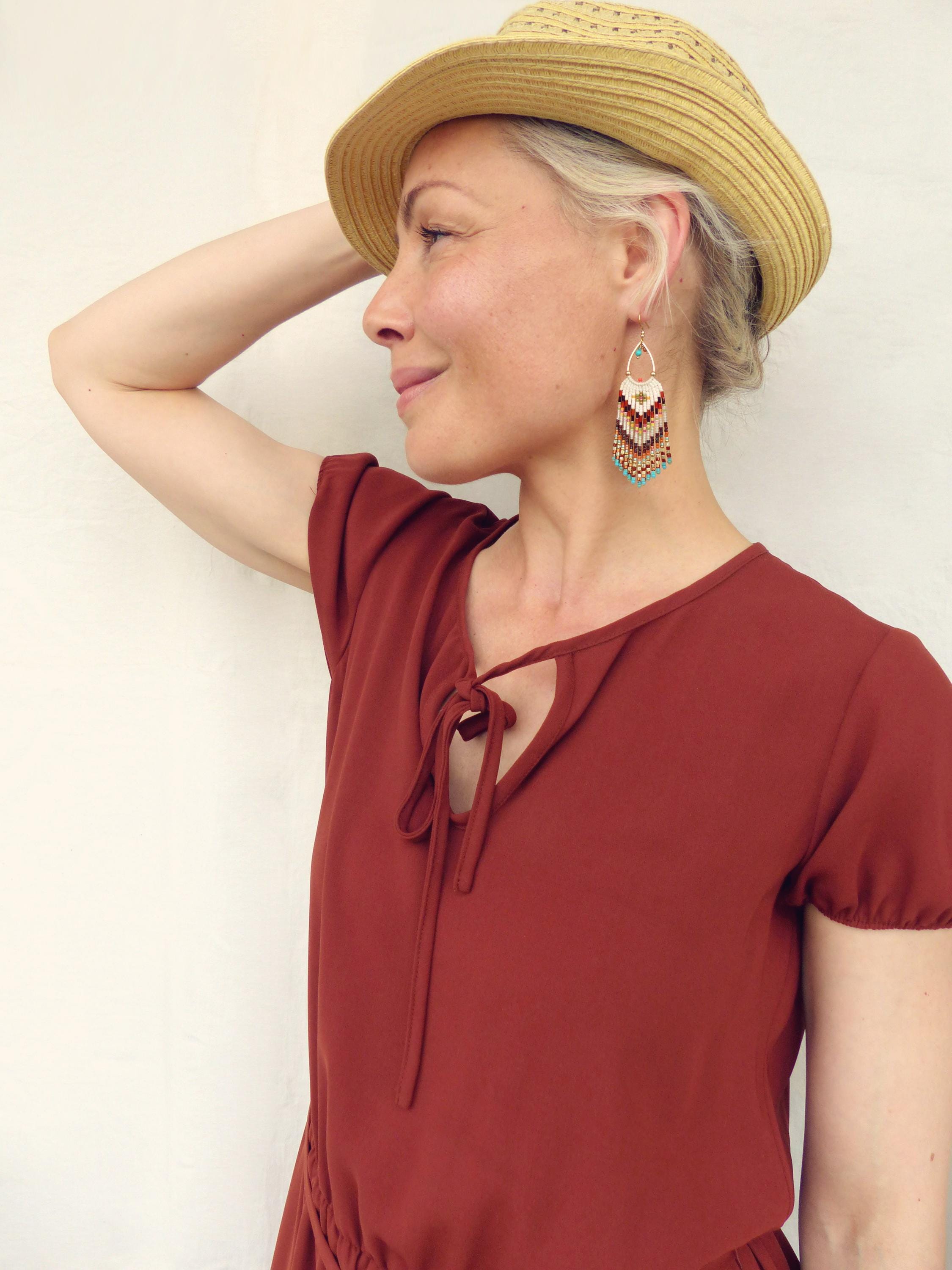 Peut inclure: Une femme porte un chapeau de paille et une robe couleur rouille, avec des boucles d'oreilles en perles blanches, marron et turquoise. Les boucles d'oreilles ont un motif géométrique. La robe a un détail de col à nouer.