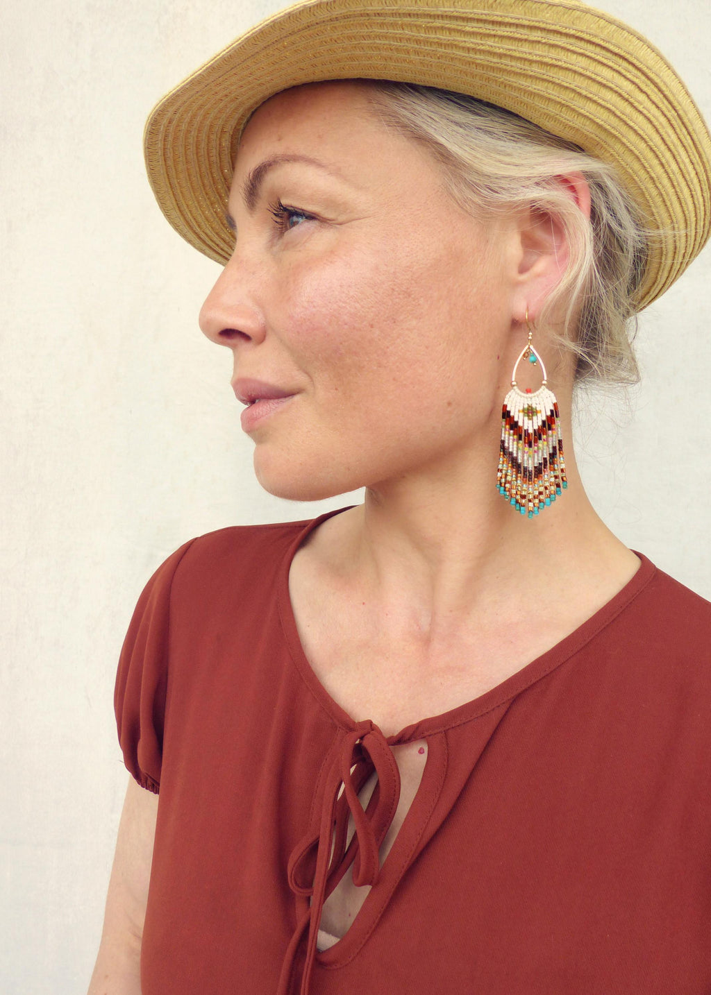 Peut inclure: Une femme porte un chapeau de paille et un chemisier couleur rouille. Elle porte des boucles d'oreilles en perles avec une forme de goutte d'eau dorée et un motif en chevrons blanc, marron, rouge, or et turquoise.