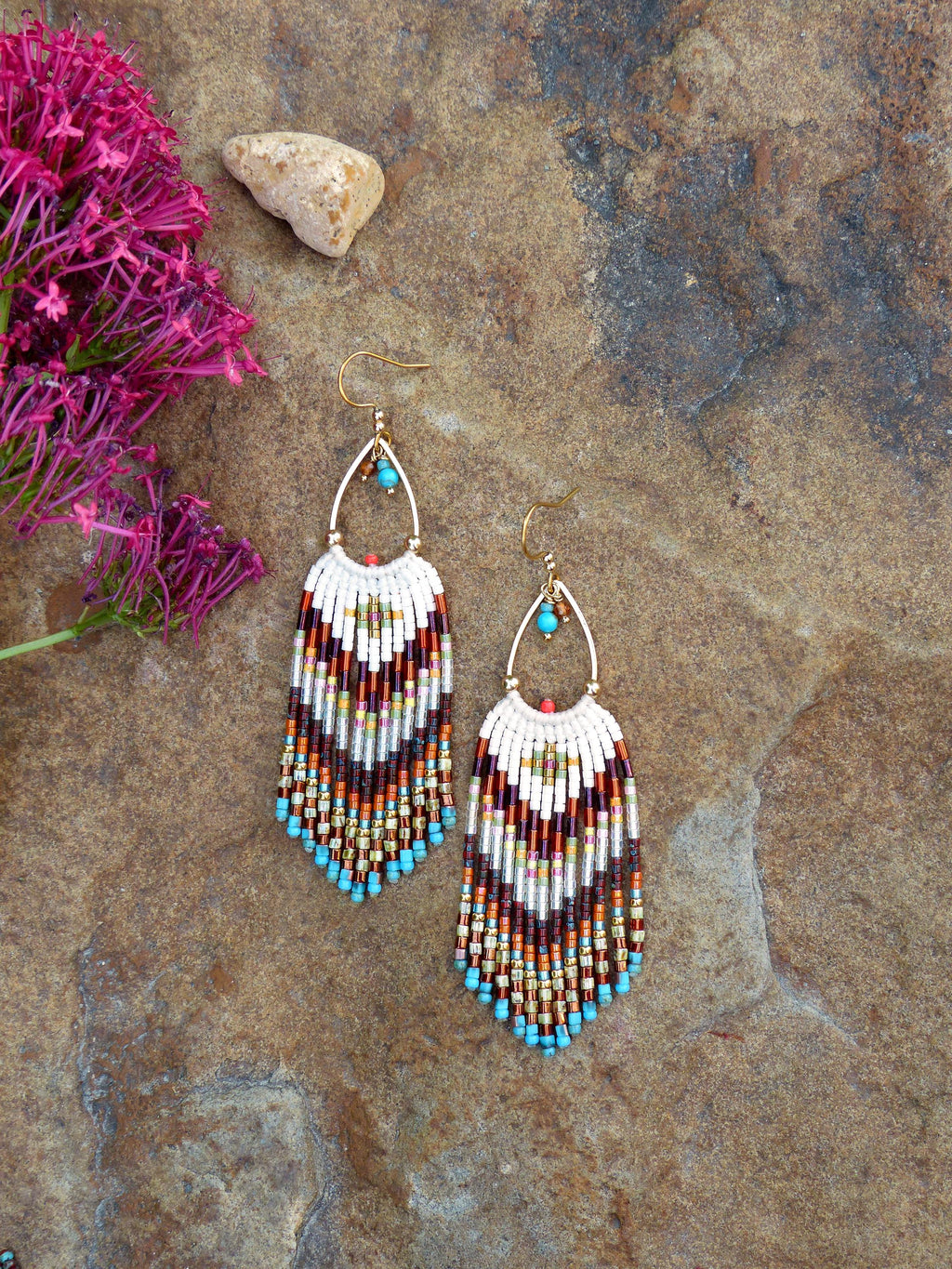 Peut inclure: Boucles d'oreilles artisanales en perles avec un cadre doré en forme de goutte d'eau. Les boucles d'oreilles présentent une partie supérieure blanche et une frange colorée de perles marron, orange et turquoise. Elles mesurent environ 7 cm de long.