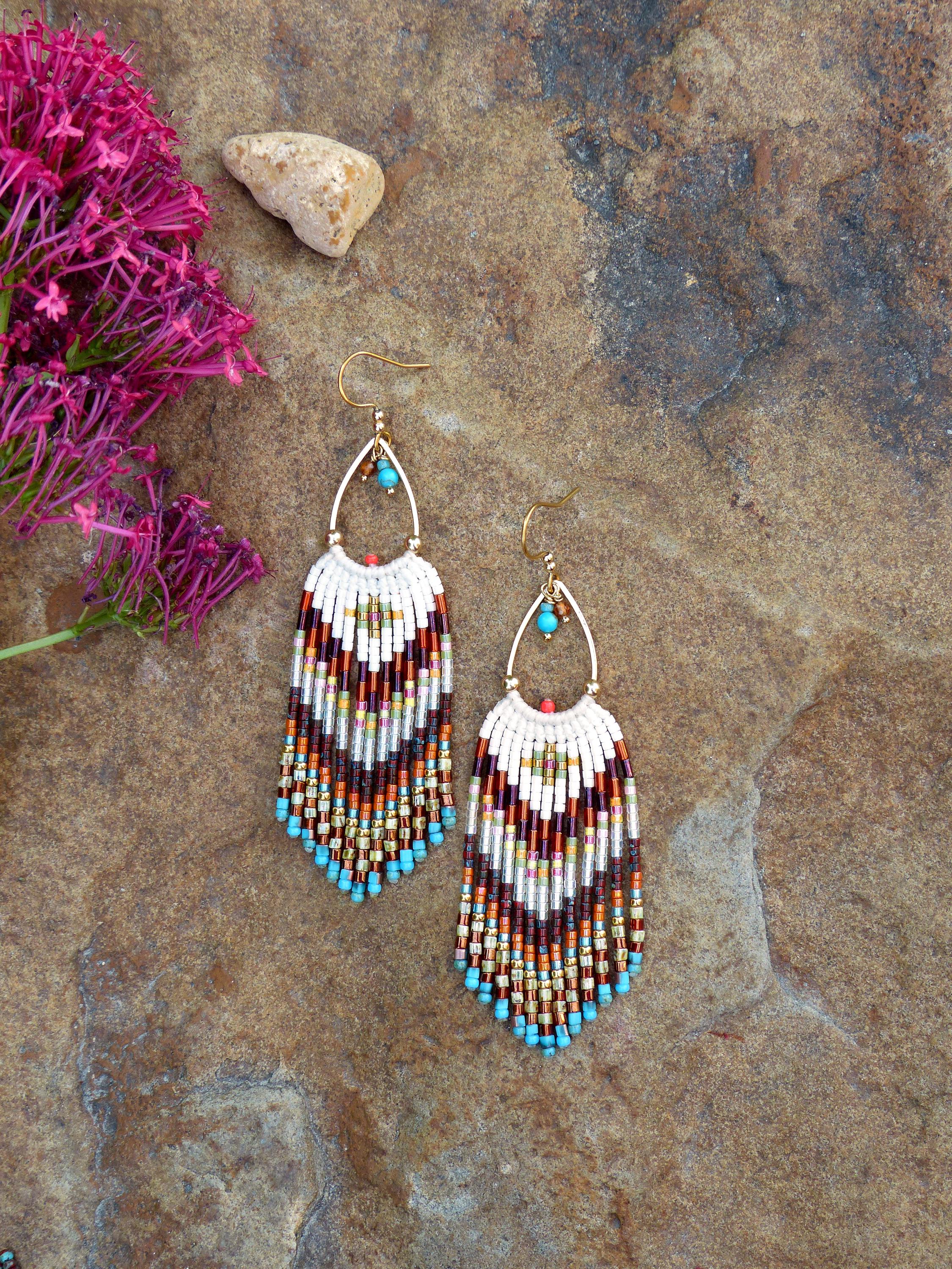 Peut inclure: Boucles d'oreilles artisanales en perles avec un cadre doré en forme de goutte d'eau. Les boucles d'oreilles présentent une partie supérieure blanche et une frange colorée de perles marron, orange et turquoise. Elles mesurent environ 7 cm de long.
