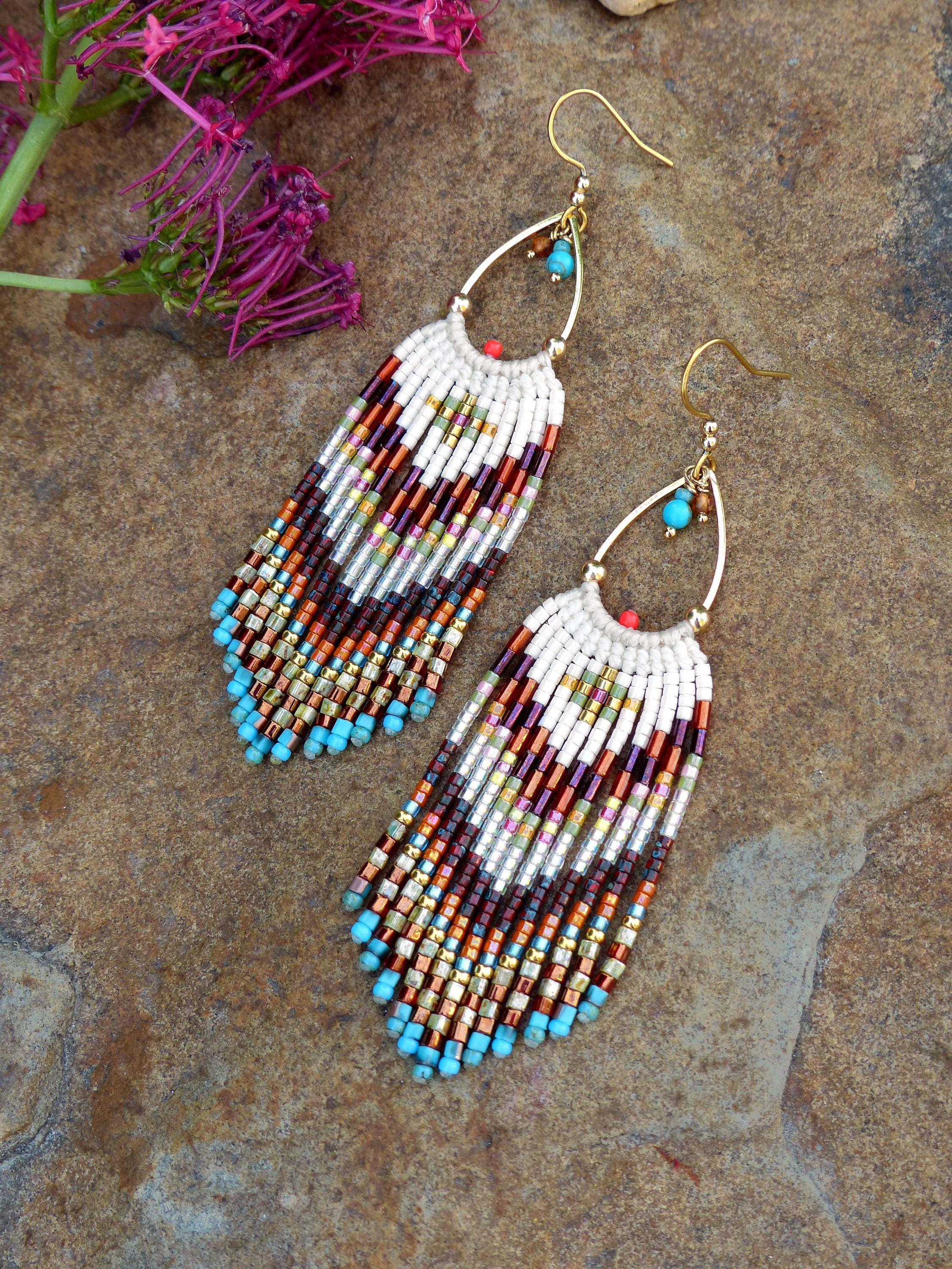 Peut inclure: Une paire de boucles d'oreilles perlées faites à la main en forme de goutte d'eau. Les boucles d'oreilles présentent une section supérieure blanche et une frange de perles colorées dans les tons de marron, orange, turquoise et or. Elles ont des crochets d'oreilles dorés et des perles turquoise.