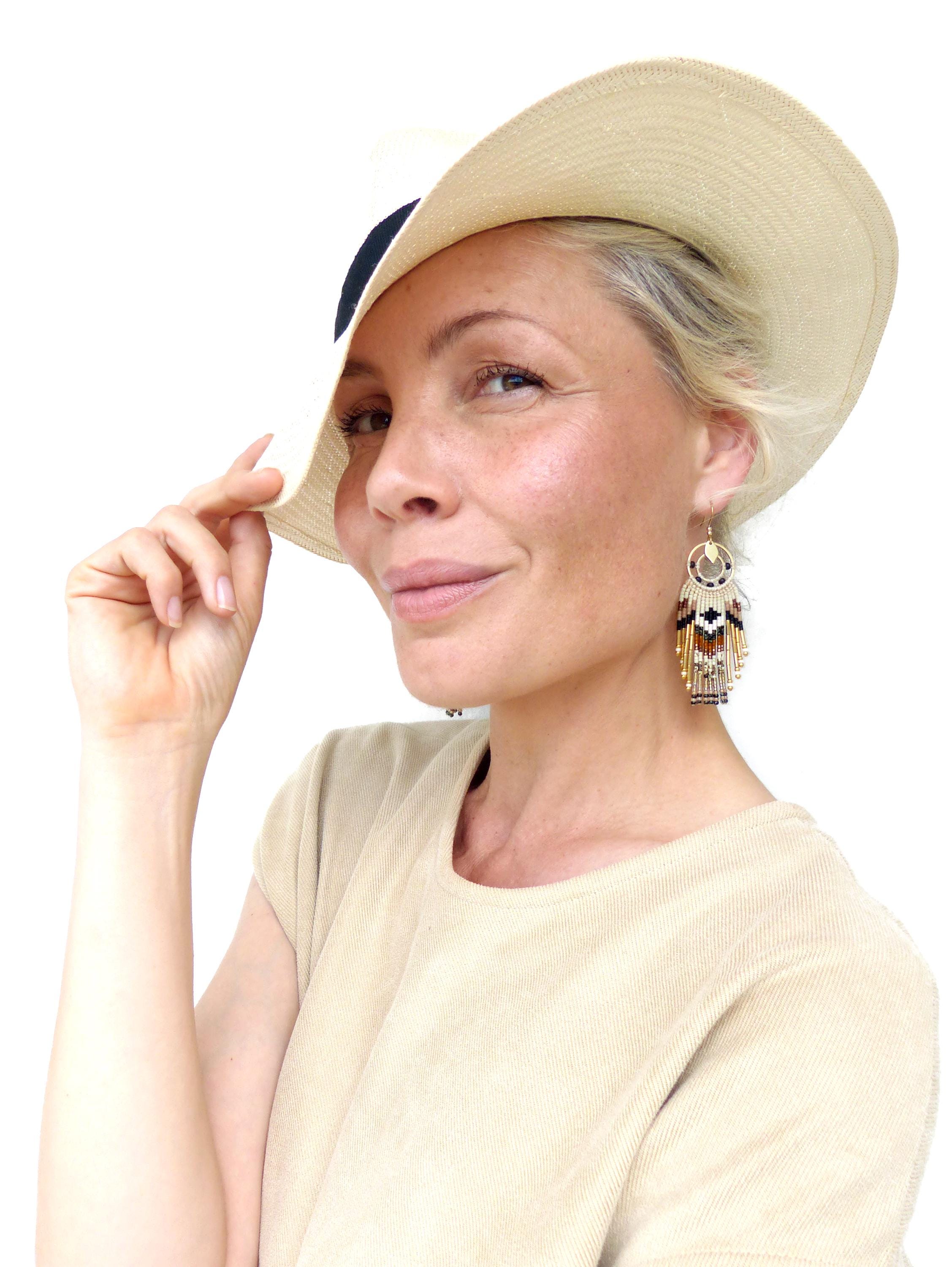 Peut inclure: Une femme porte un chapeau de soleil beige avec une bande noire et des boucles d'oreilles perlées. Les boucles d'oreilles présentent un motif circulaire avec des motifs géométriques noirs, blancs et dorés, avec des franges perlées pendantes.