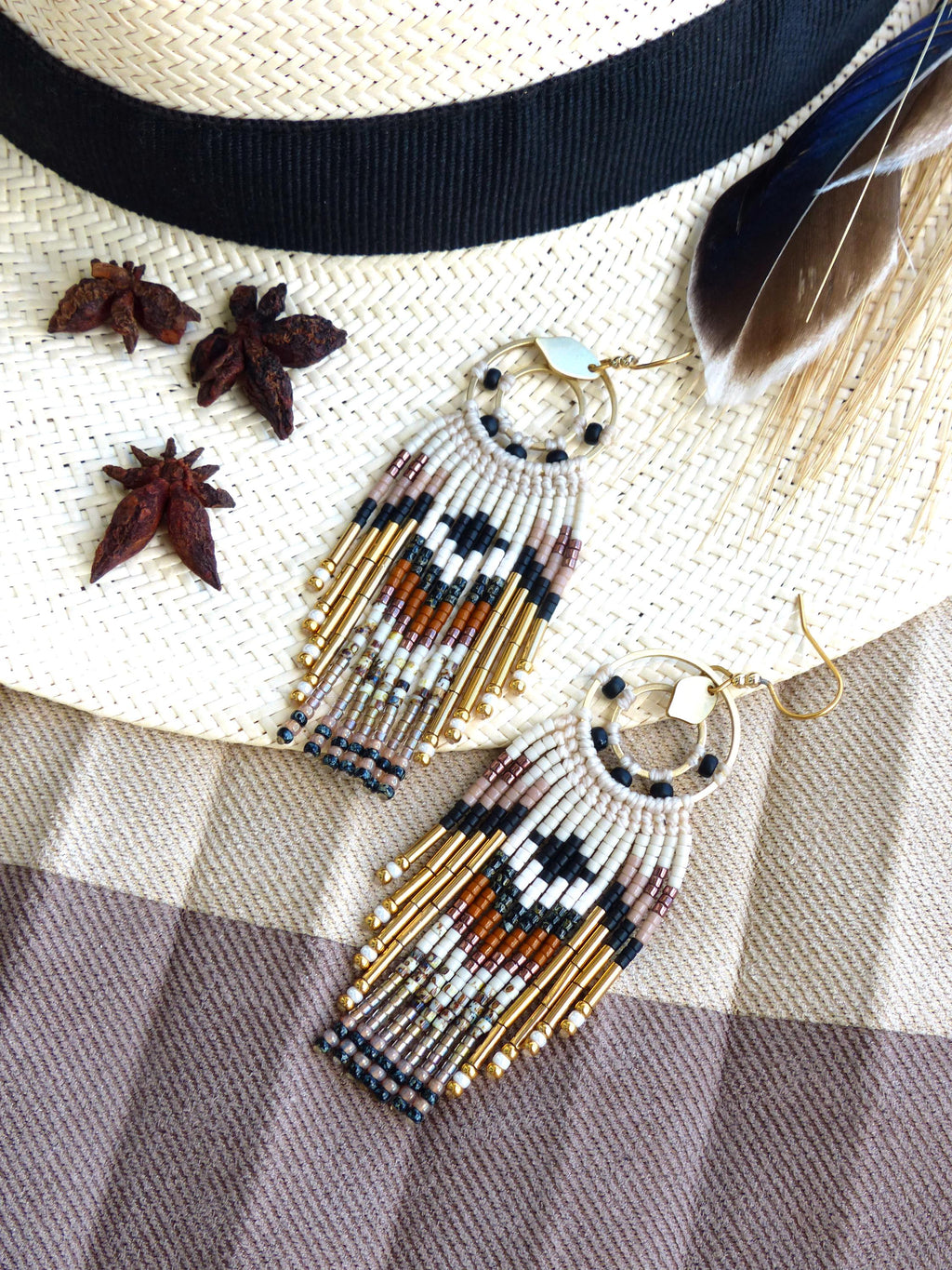 Peut inclure: Boucles d'oreilles faites à la main avec un motif géométrique en crème, noir, marron et or. Les boucles d'oreilles ont un dessus circulaire et une frange de perles. Elles sont présentées sur un chapeau de paille.