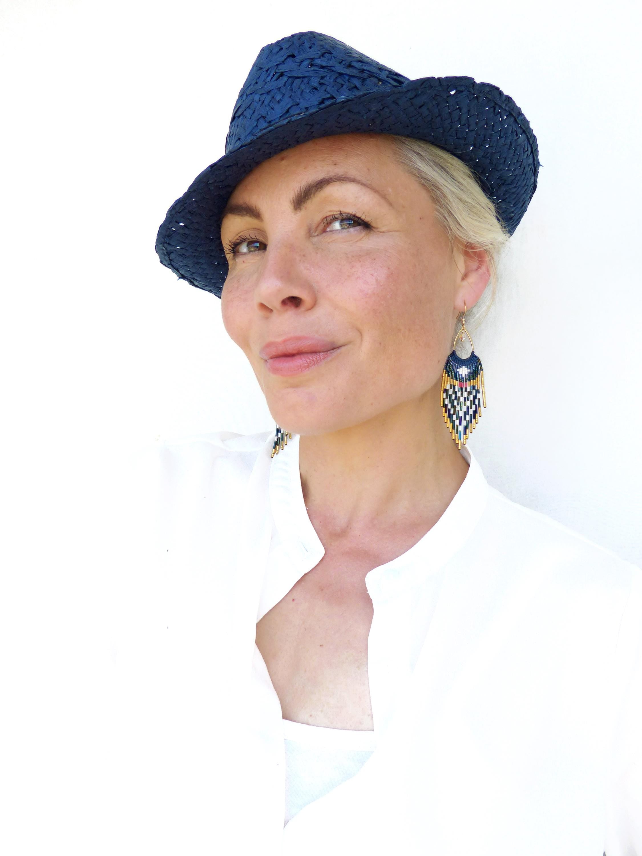 Peut inclure: Un chapeau fedora bleu marine avec une texture tissée. L'image présente également des boucles d'oreilles perlées avec un motif géométrique bleu, blanc, jaune et noir. La personne porte une chemise blanche.