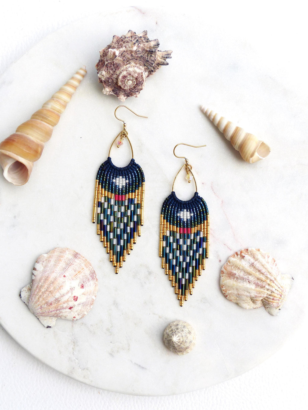 Peut inclure: Boucles d'oreilles perlées faites à la main avec une partie supérieure bleu marine et un motif géométrique blanc, vert, rouge et or. Les boucles d'oreilles sont présentées avec des coquillages sur une surface en marbre blanc, créant un thème côtier.