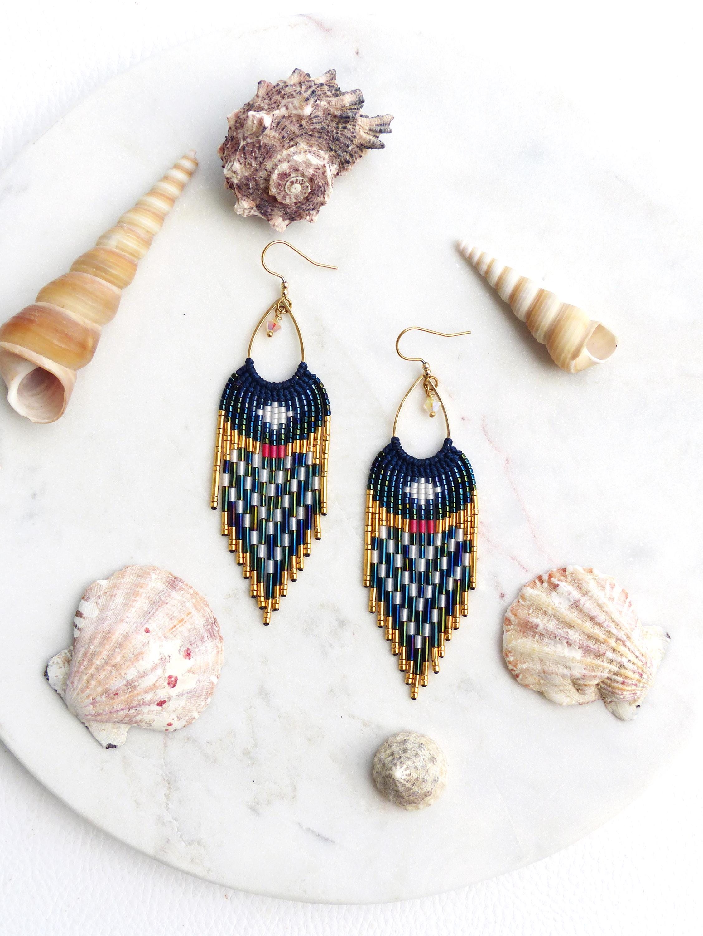 Peut inclure: Boucles d'oreilles perlées faites à la main avec une partie supérieure bleu marine et un motif géométrique blanc, vert, rouge et or. Les boucles d'oreilles sont présentées avec des coquillages sur une surface en marbre blanc, créant un thème côtier.