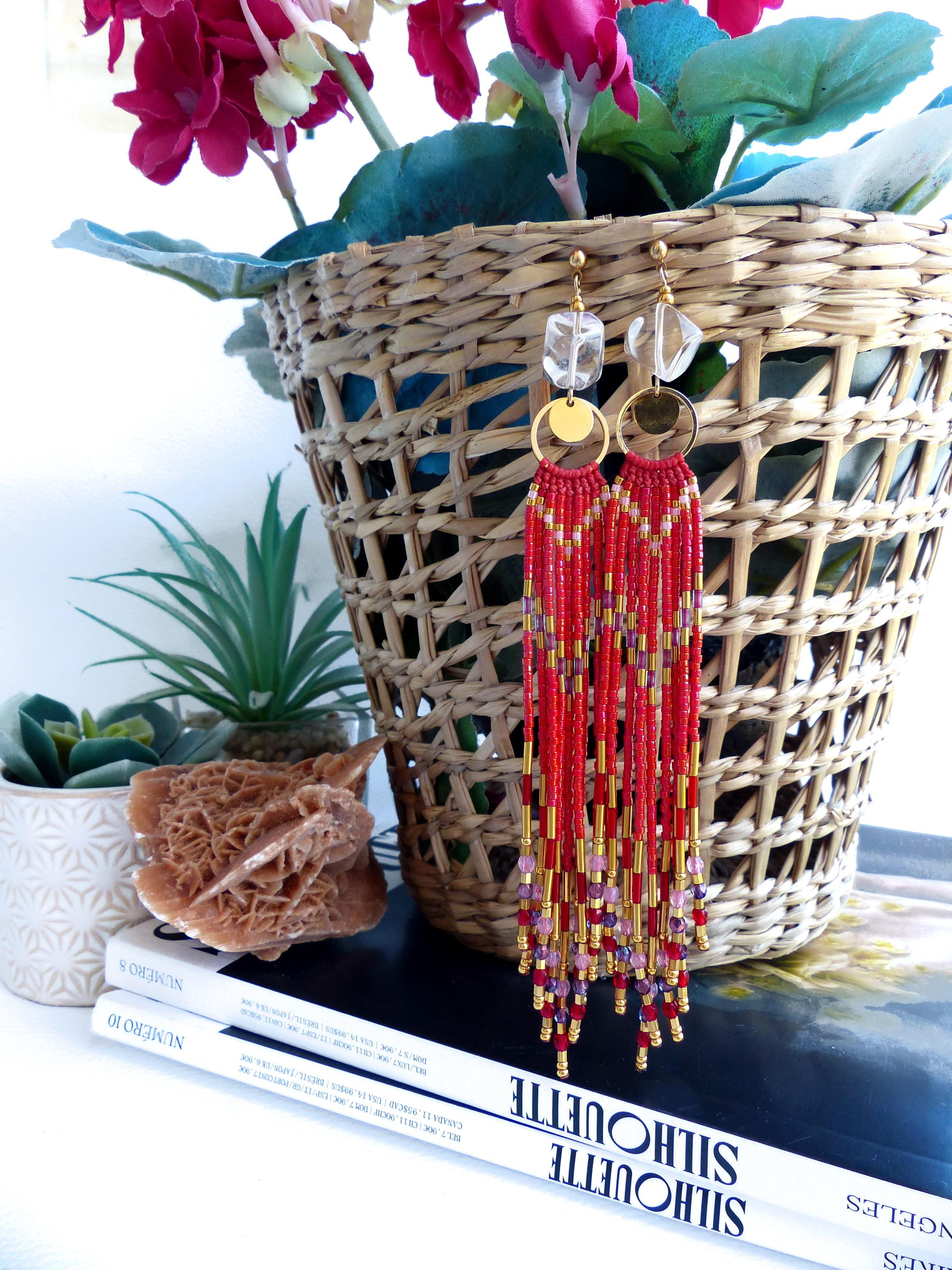Peut inclure: Longues boucles d'oreilles rouges à perles avec des accents dorés et des dessus en cristal transparent. Les boucles d'oreilles présentent un motif de franges avec un mélange de perles rouges, dorées et violettes. Elles sont devant un panier en osier avec des fleurs.