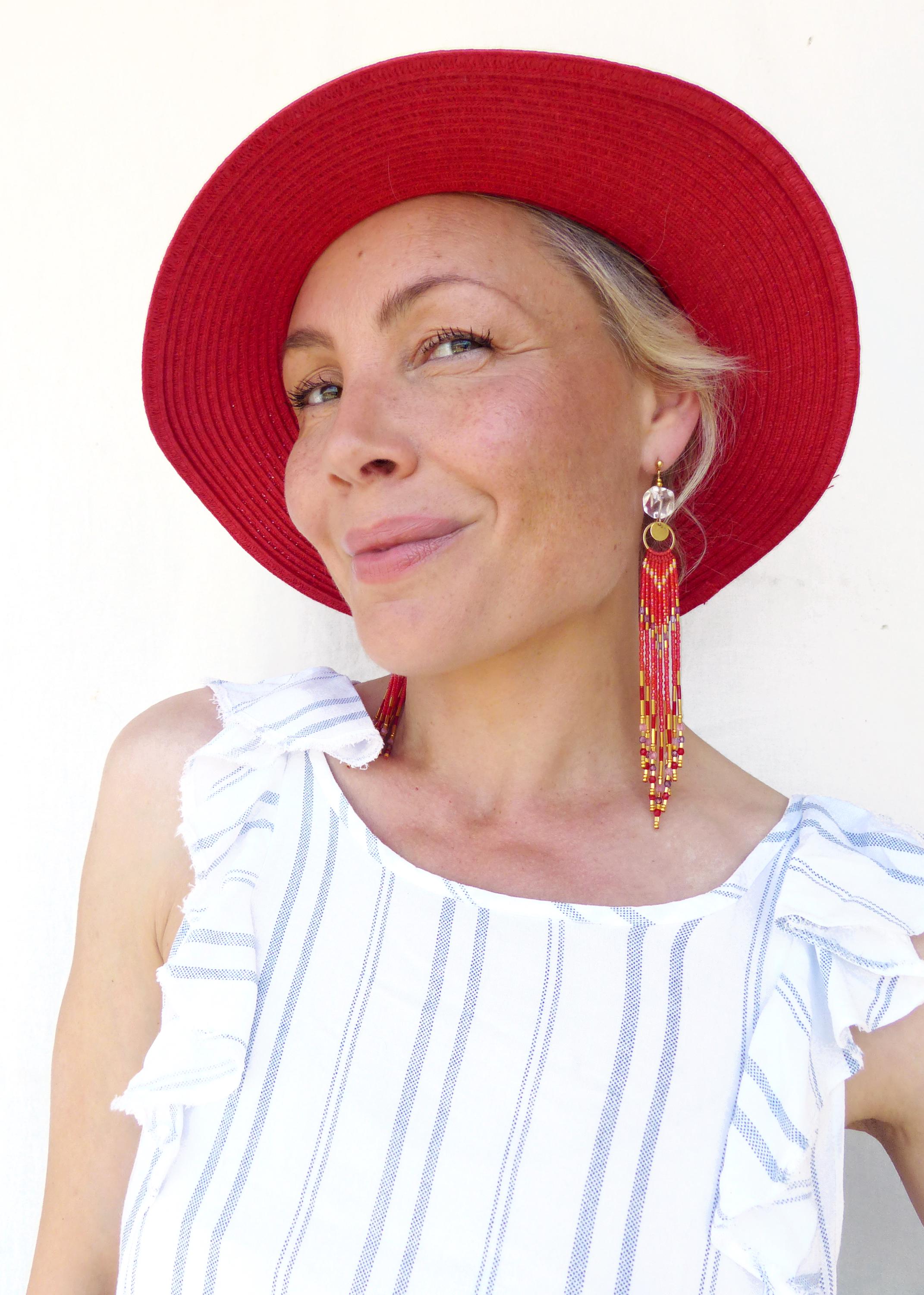 Peut inclure: Une femme porte un chapeau de soleil rouge et de longues boucles d'oreilles en perles. Les boucles d'oreilles sont principalement rouges, avec des accents jaunes et dorés. Elle porte un haut blanc à rayures bleues.