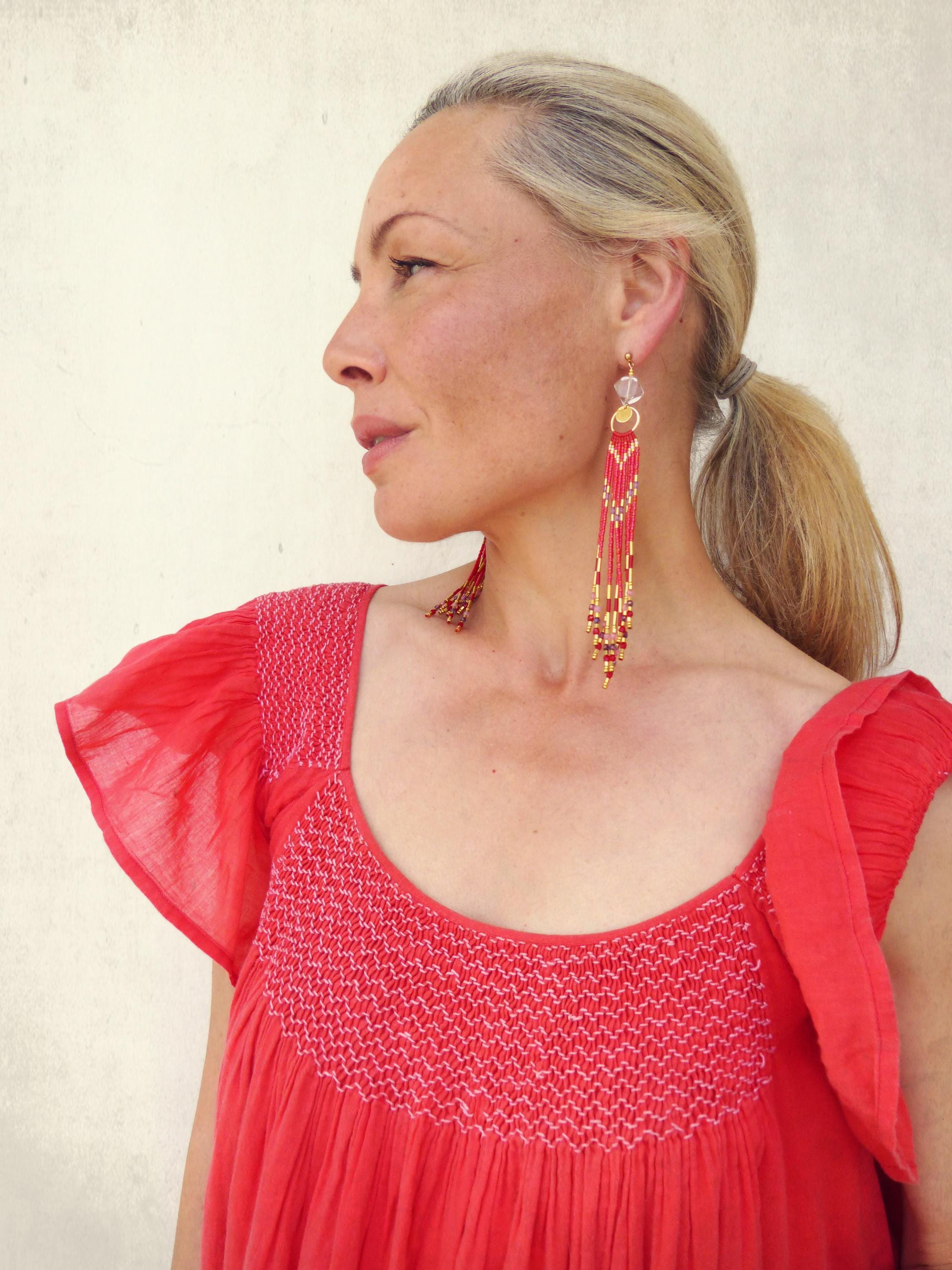 Peut inclure: Une femme porte de longues boucles d'oreilles perlées et une robe rouge. Les boucles d'oreilles sont principalement rouges avec des accents dorés et blancs, avec un design en couches. La robe a un col carré et des manches à volants.