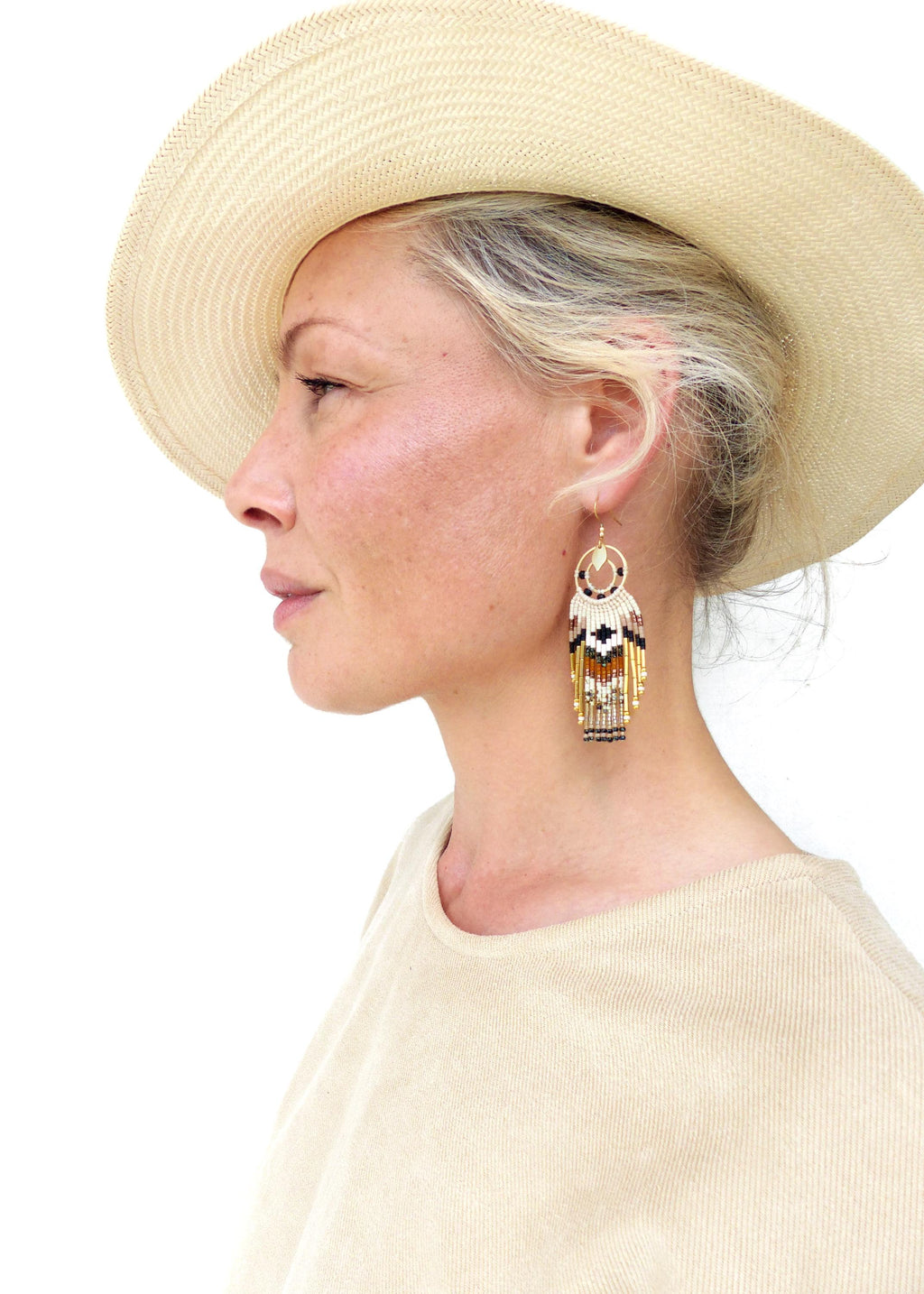 Peut inclure: Une femme porte un chapeau beige et un haut beige. Elle porte des boucles d'oreilles dorées avec un motif de perles noires, blanches, marron et dorées. Les boucles d'oreilles ont une frange de perles.