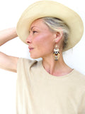 Peut inclure: Boucles d'oreilles pendantes avec un motif géométrique en perles noires, blanches et dorées. Elles sont portées avec un chapeau et un haut beiges.