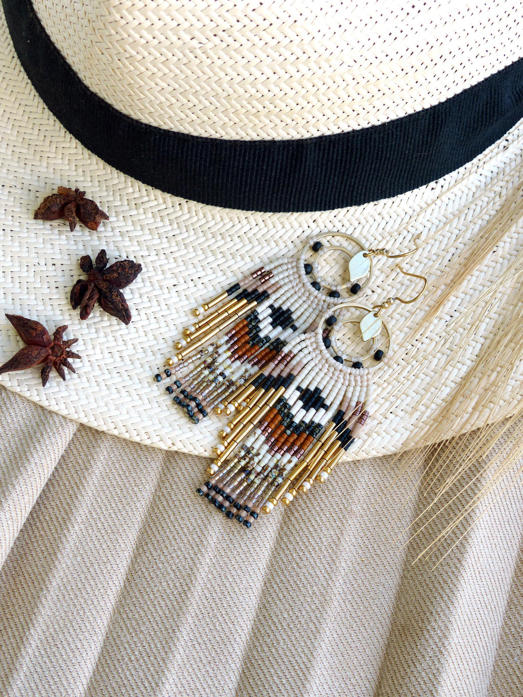 Peut inclure: Boucles d'oreilles perlées faites à la main avec un motif géométrique noir, blanc, marron et or. Les boucles d'oreilles ont un design à franges et sont présentées sur un chapeau de paille avec un ruban noir. De l'anis étoilé est dispersé.