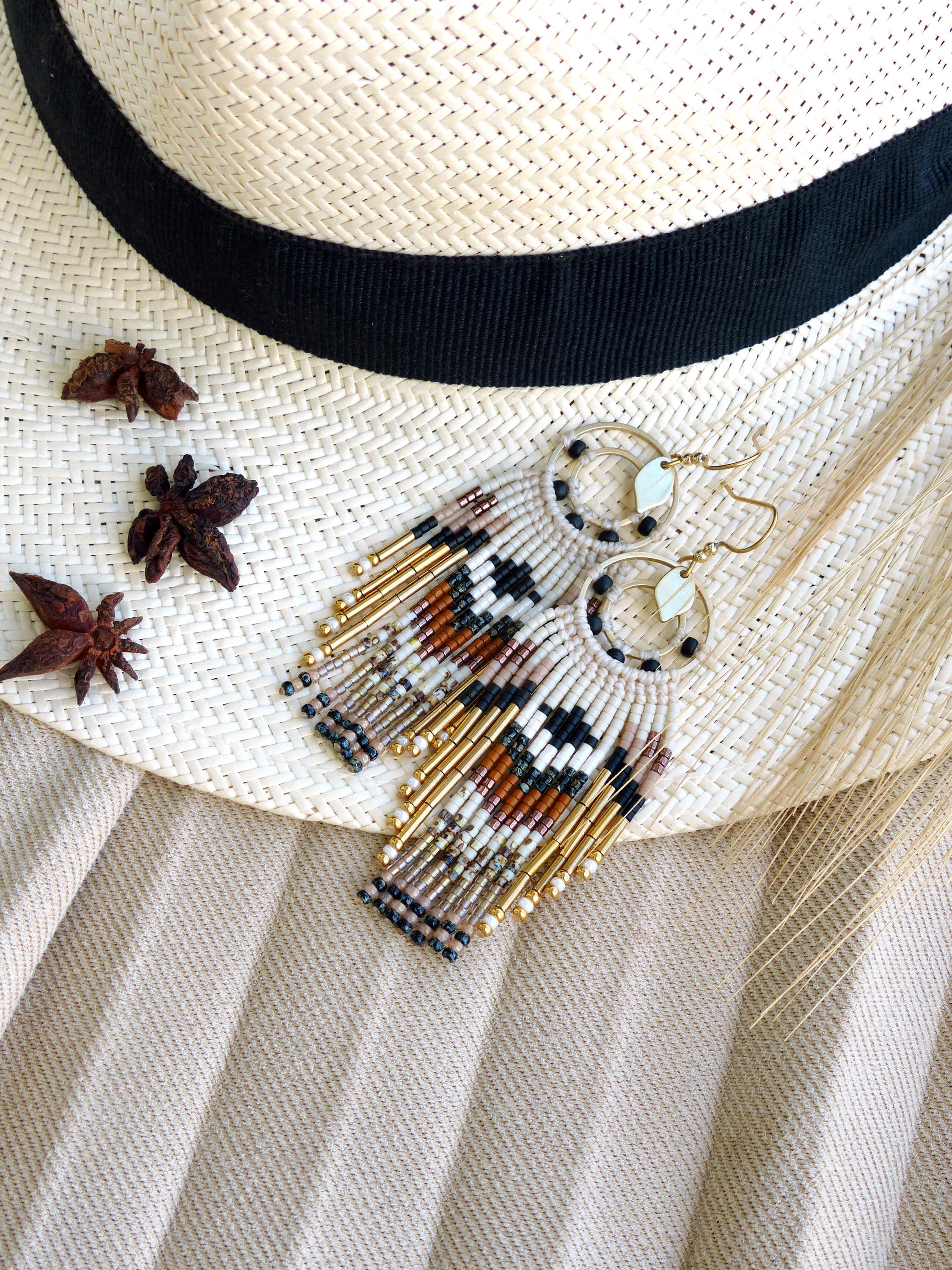 Peut inclure: Boucles d'oreilles perlées faites à la main avec un motif géométrique noir, blanc, marron et or. Les boucles d'oreilles ont un design à franges et sont présentées sur un chapeau de paille avec un ruban noir. De l'anis étoilé est dispersé.