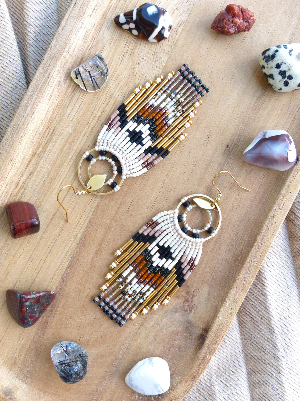Peut inclure: Boucles d'oreilles faites à la main avec un motif géométrique en blanc, noir, marron et or. Les boucles d'oreilles ont une frange de perles dorées et sont présentées sur un plateau en bois avec diverses pierres précieuses.