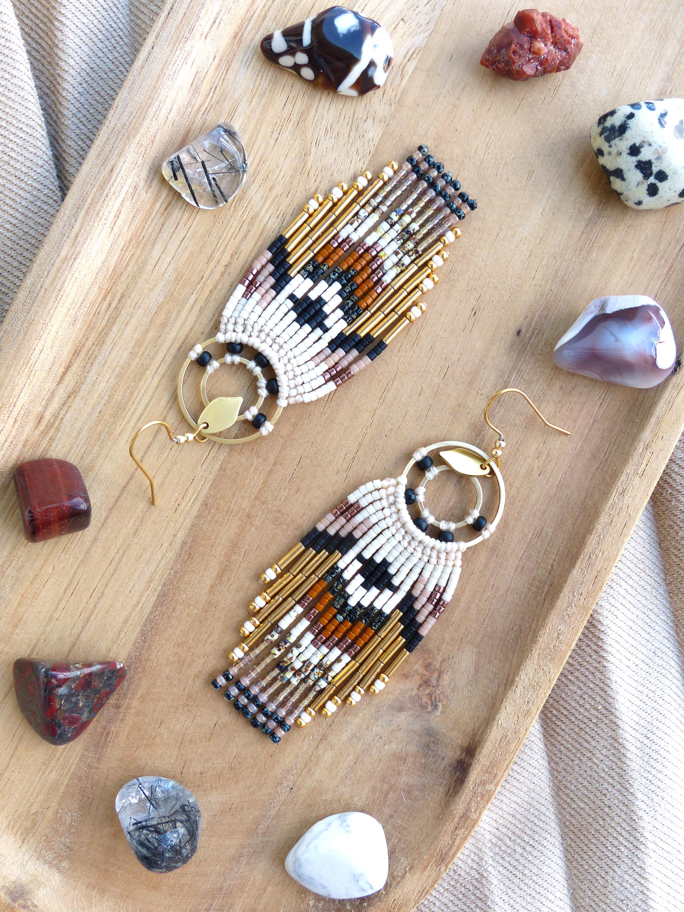 Peut inclure: Boucles d'oreilles faites à la main avec un motif géométrique en blanc, noir, marron et or. Les boucles d'oreilles ont une frange de perles dorées et sont présentées sur un plateau en bois avec diverses pierres précieuses.