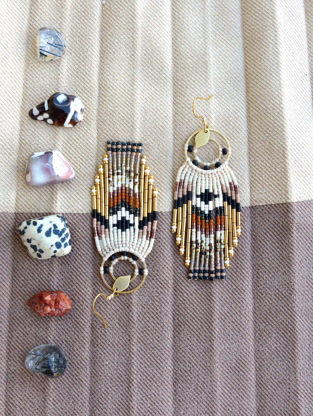 Peut inclure: Boucles d'oreilles faites à la main avec un motif géométrique en noir, blanc, marron et or. Les boucles d'oreilles ont un aspect de frange et sont présentées avec diverses pierres colorées sur un fond en tissu rayé.
