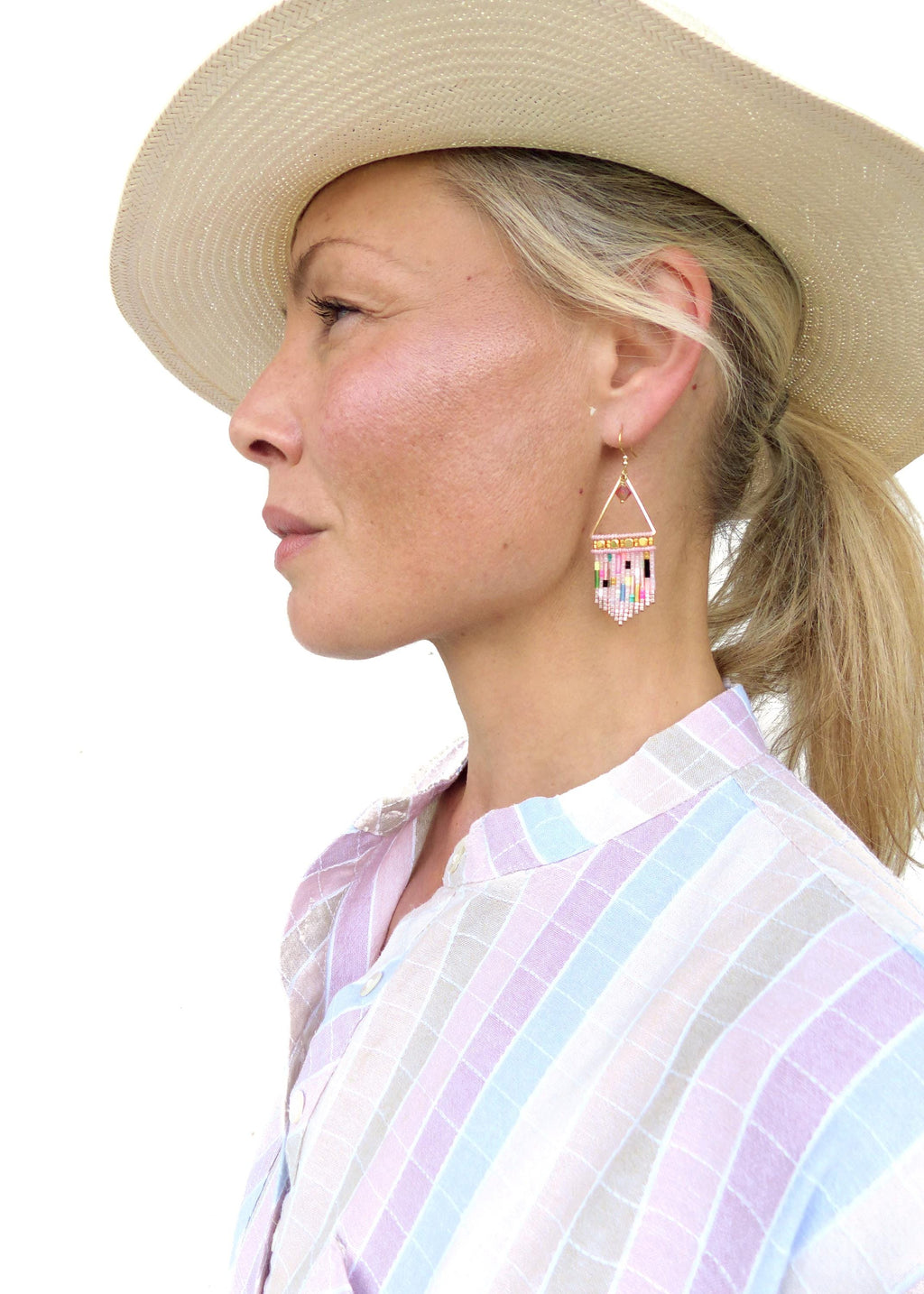 Peut inclure: Boucles d'oreilles triangulaires dorées avec un motif de perles roses, blanches, bleues et vertes. Les boucles d'oreilles sont portées par une personne portant un chapeau de paille et une chemise à carreaux pastel.