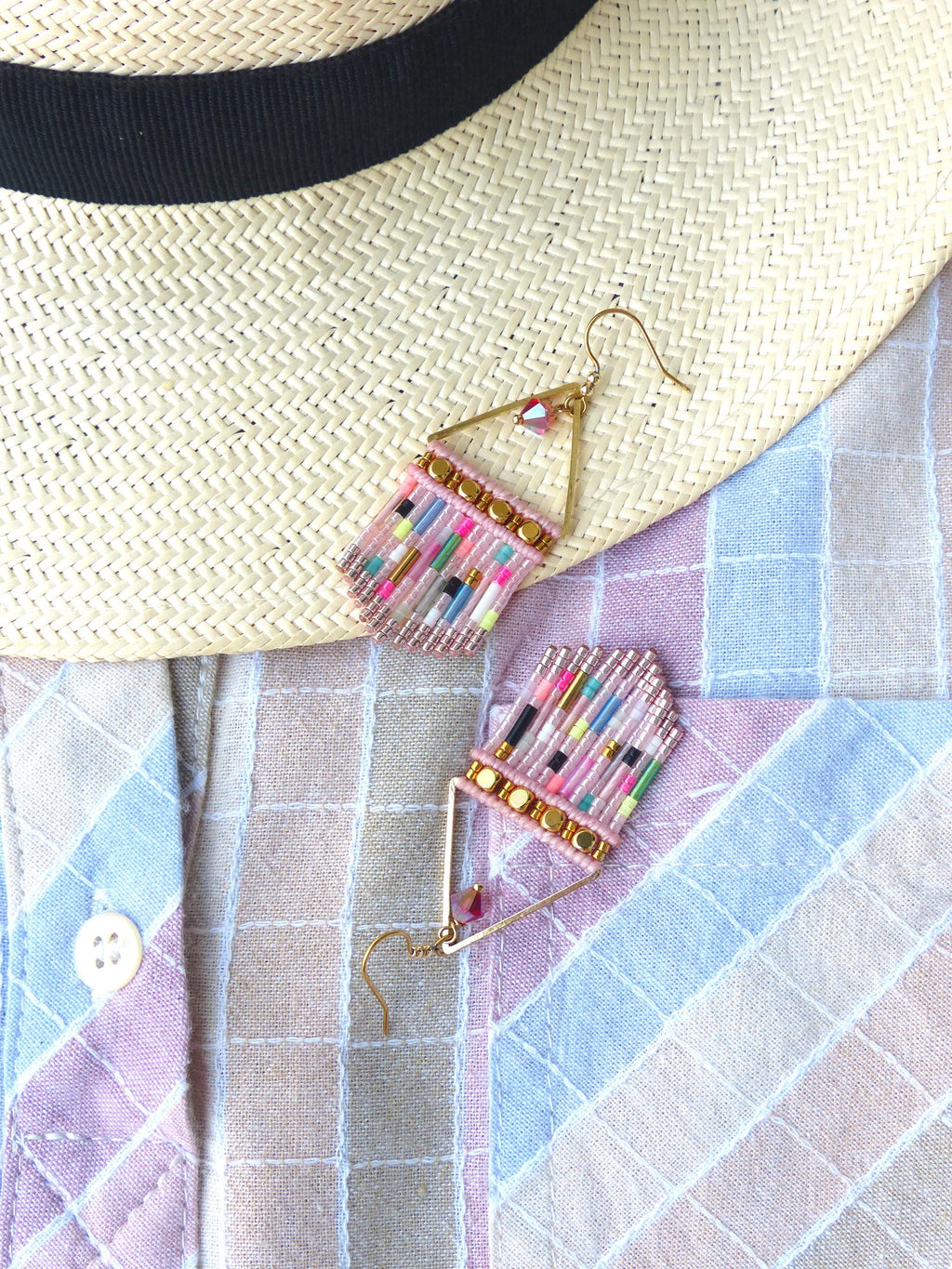 Peut inclure: Boucles d'oreilles en perles de forme géométrique. Elles présentent une base rectangulaire rose ornée de perles colorées et d'éléments métalliques dorés. Un chapeau de paille à bande noire et une chemise à carreaux pastel sont en arrière-plan.